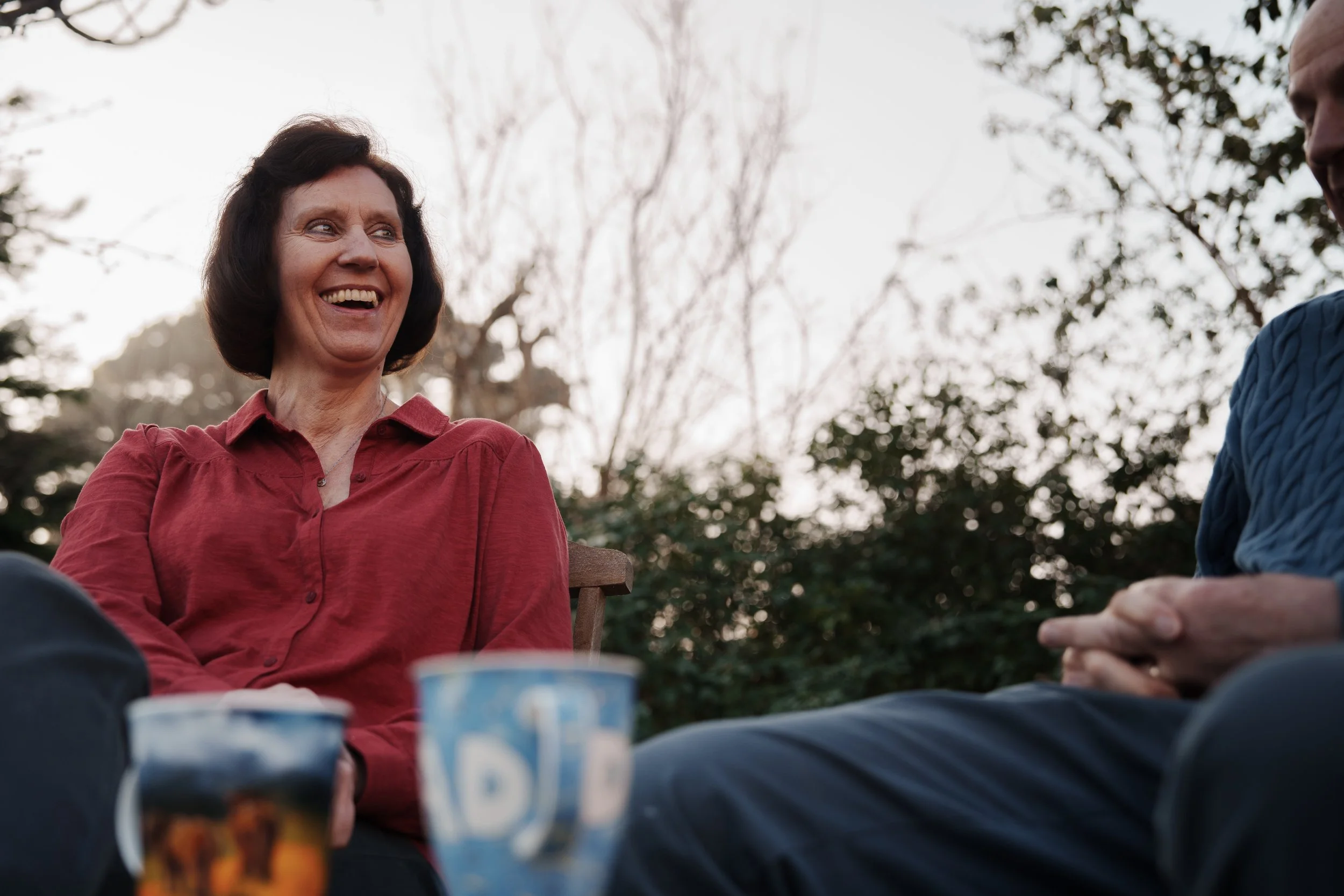 A woman with dark hair wearing a red shirt smiling and talking to a man with a blue shirt sitting outdoors during daytime.