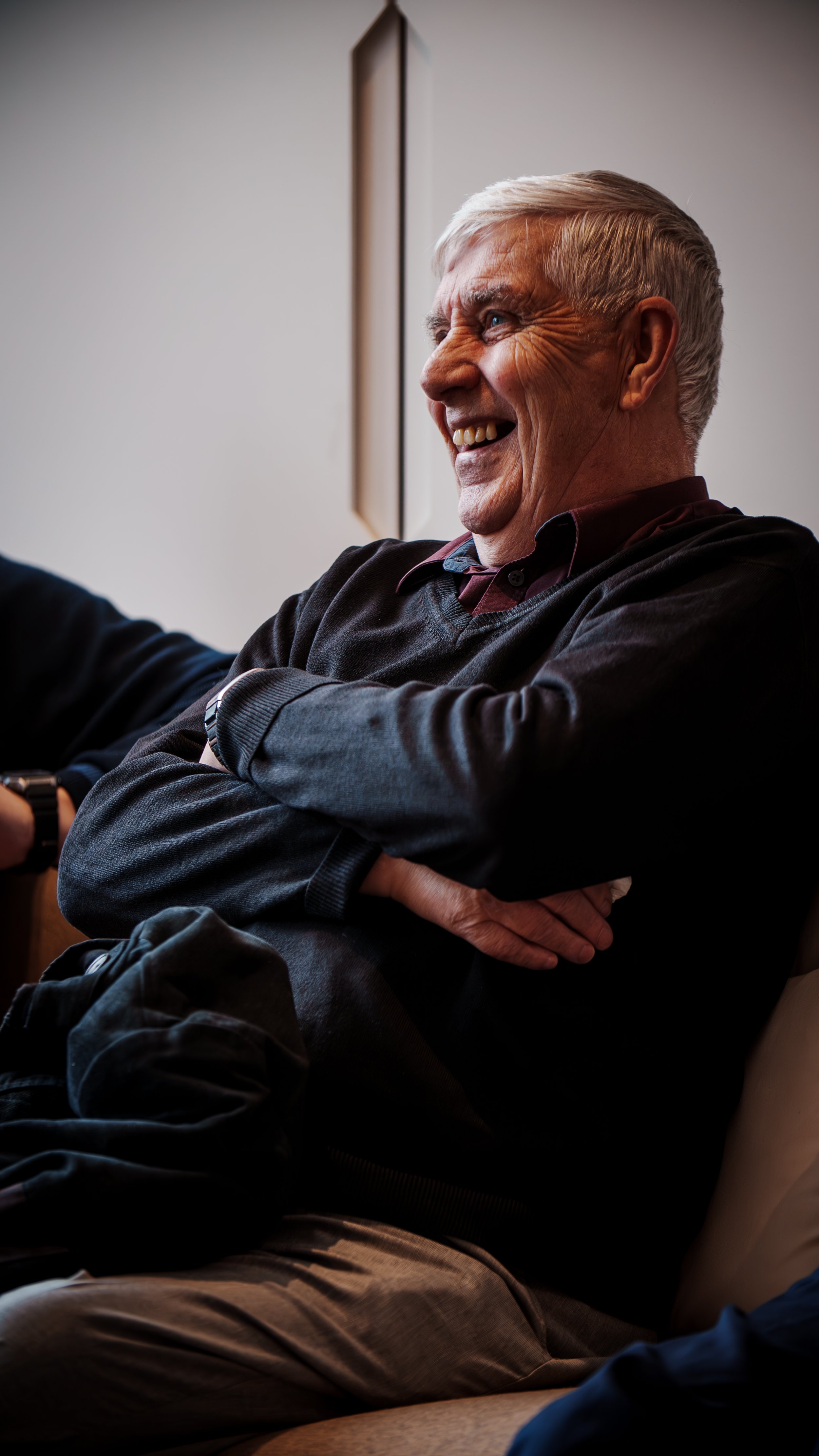 Older man with gray hair and a joyful expression, sitting with arms crossed, indoors.
