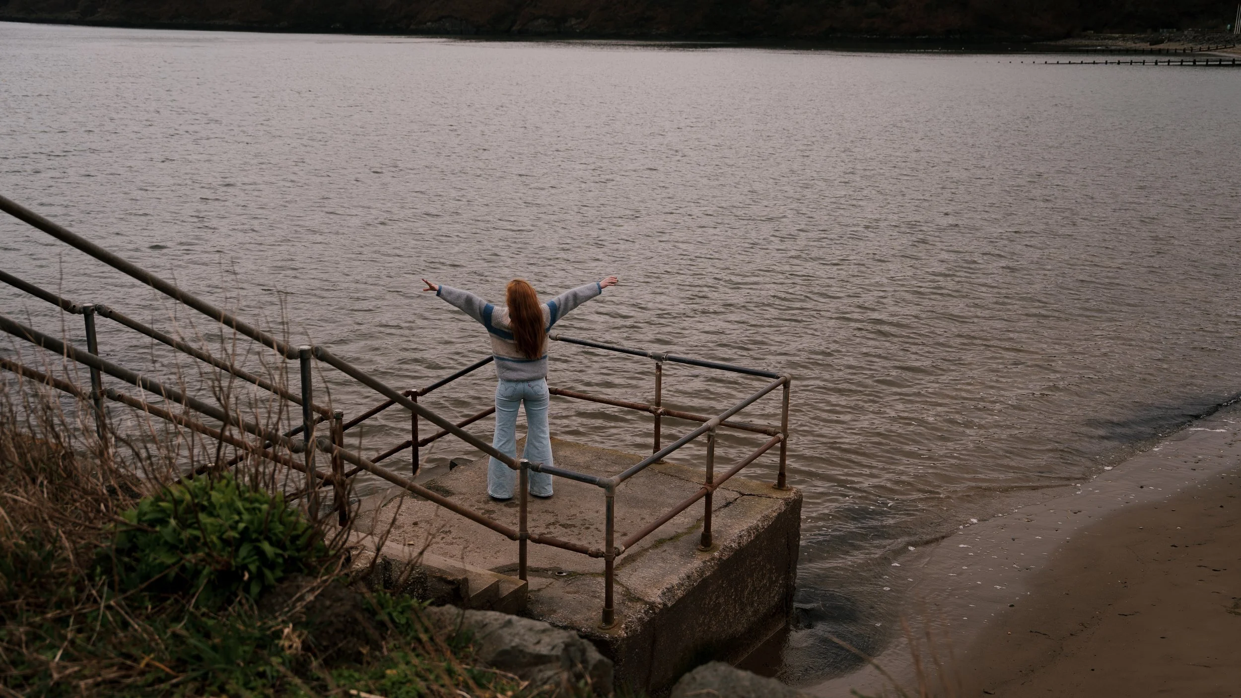 Woman with long red hair in a striped sweater and light jeans standing on a concrete platform with metal railings by the water, facing away with arms outstretched.