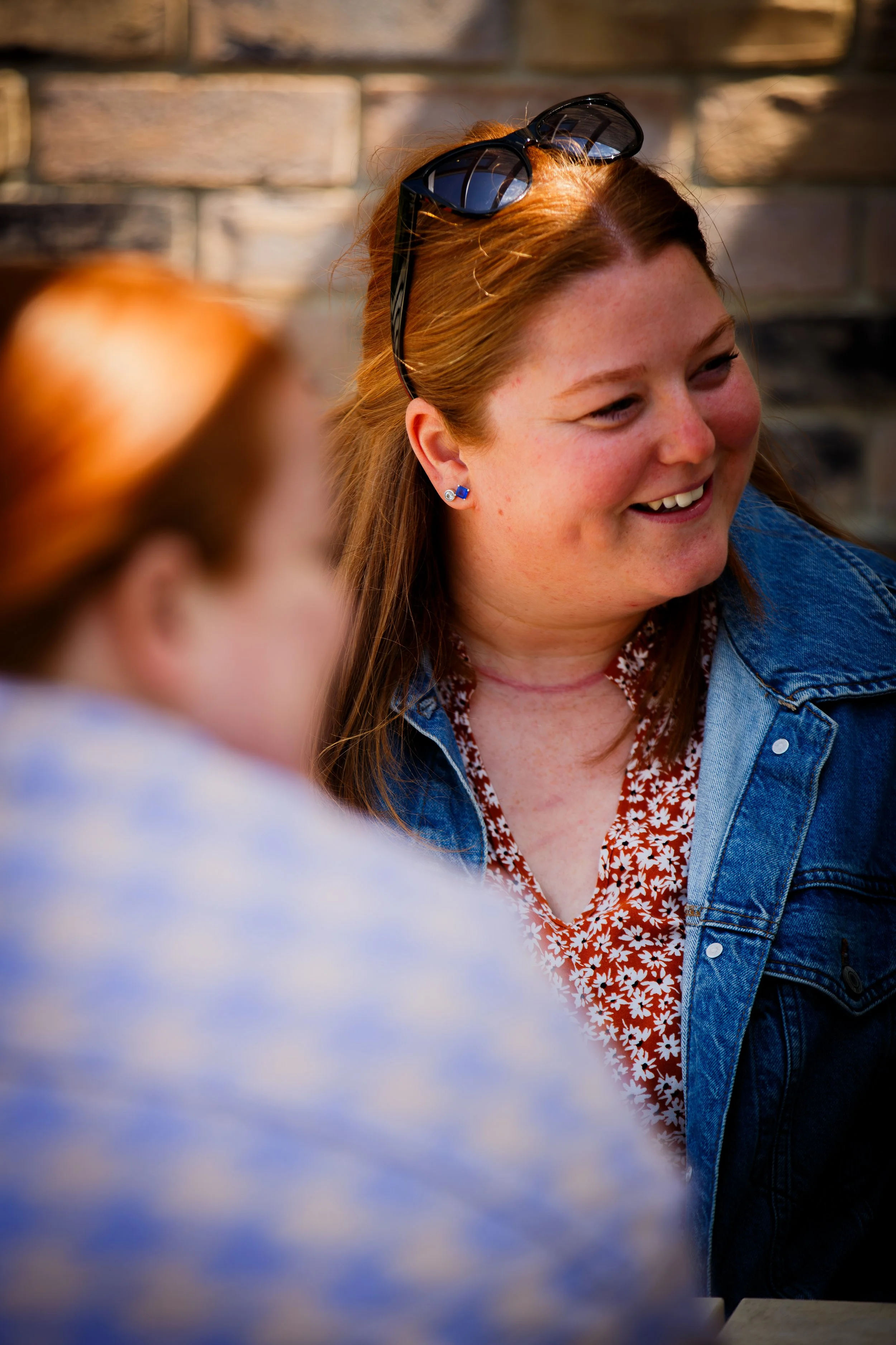 A smiling woman with sunglasses on her head, wearing a denim jacket and a floral shirt, sitting outdoors by a brick wall. A woman is blurred in the foreground.