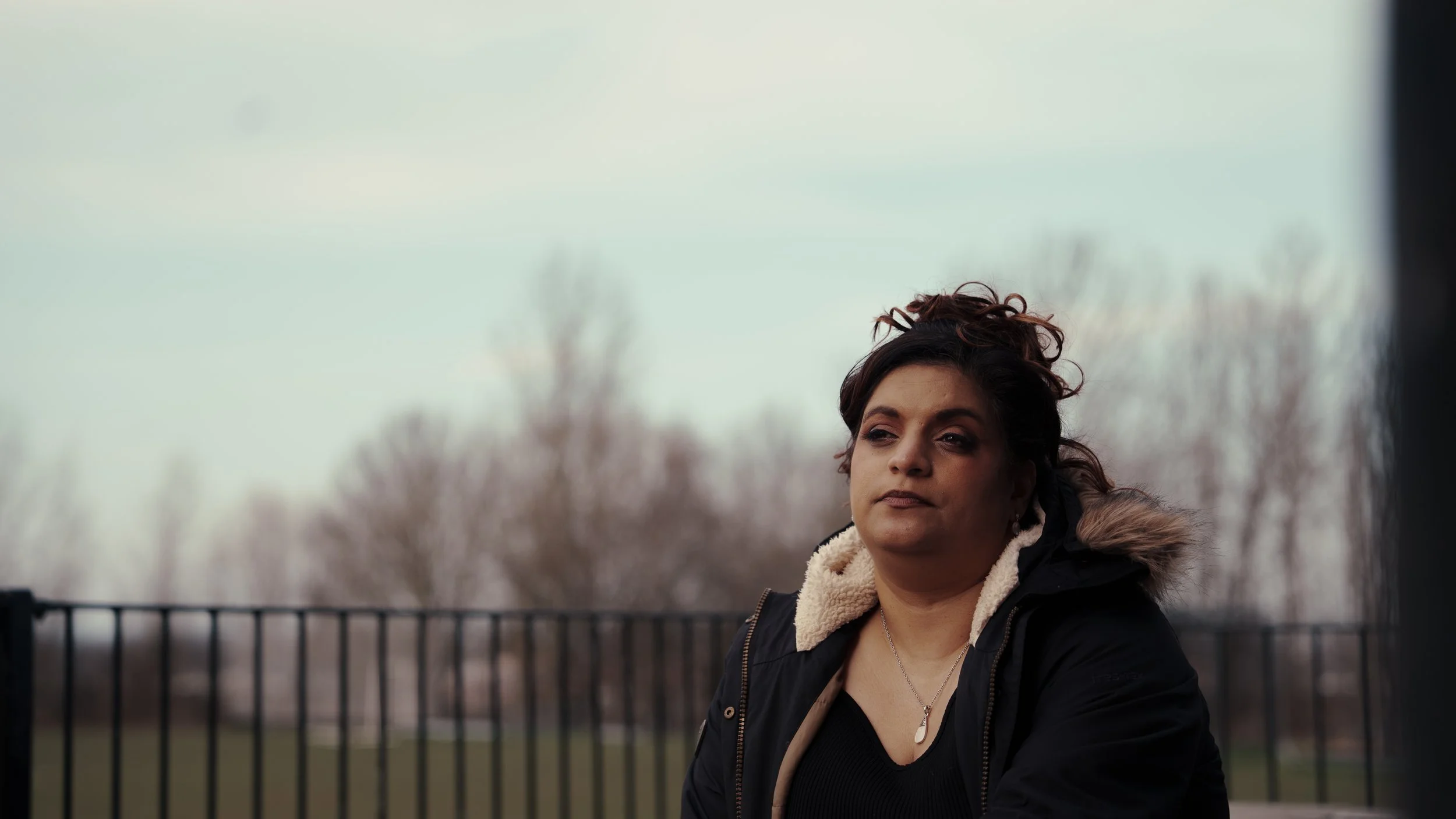 A woman with dark hair outdoors, wearing a black jacket with a serious expression.