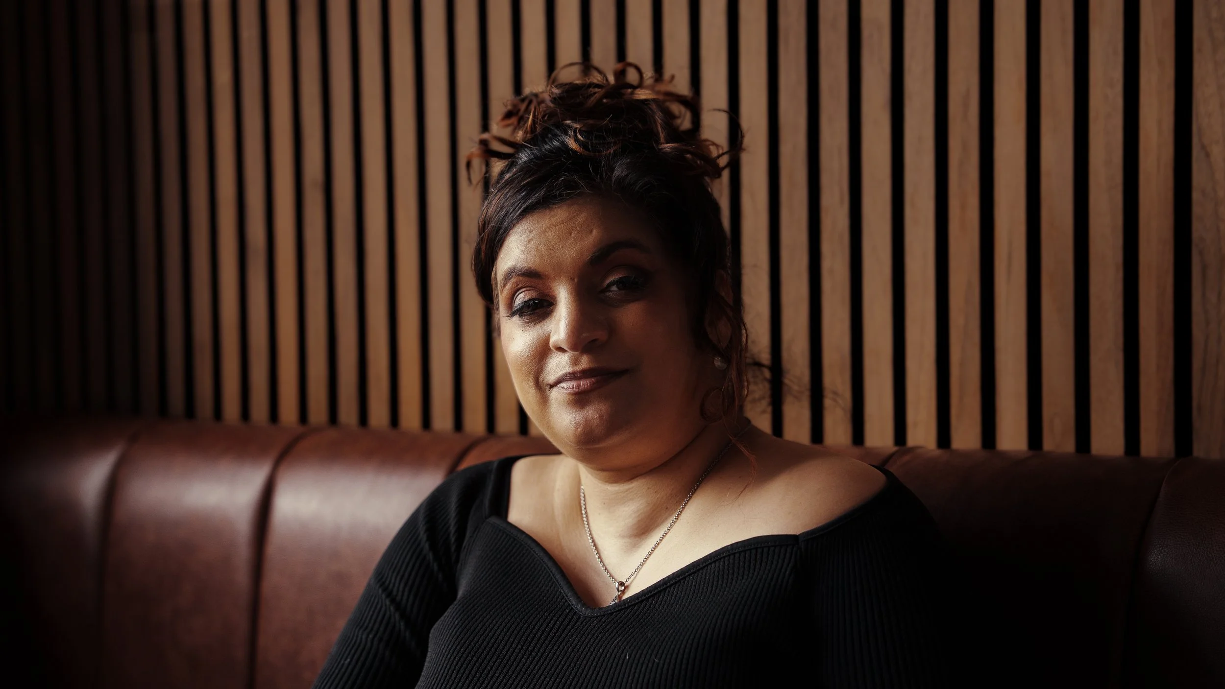 A woman with curly hair and a black top sitting on a brown booth in a cafe.