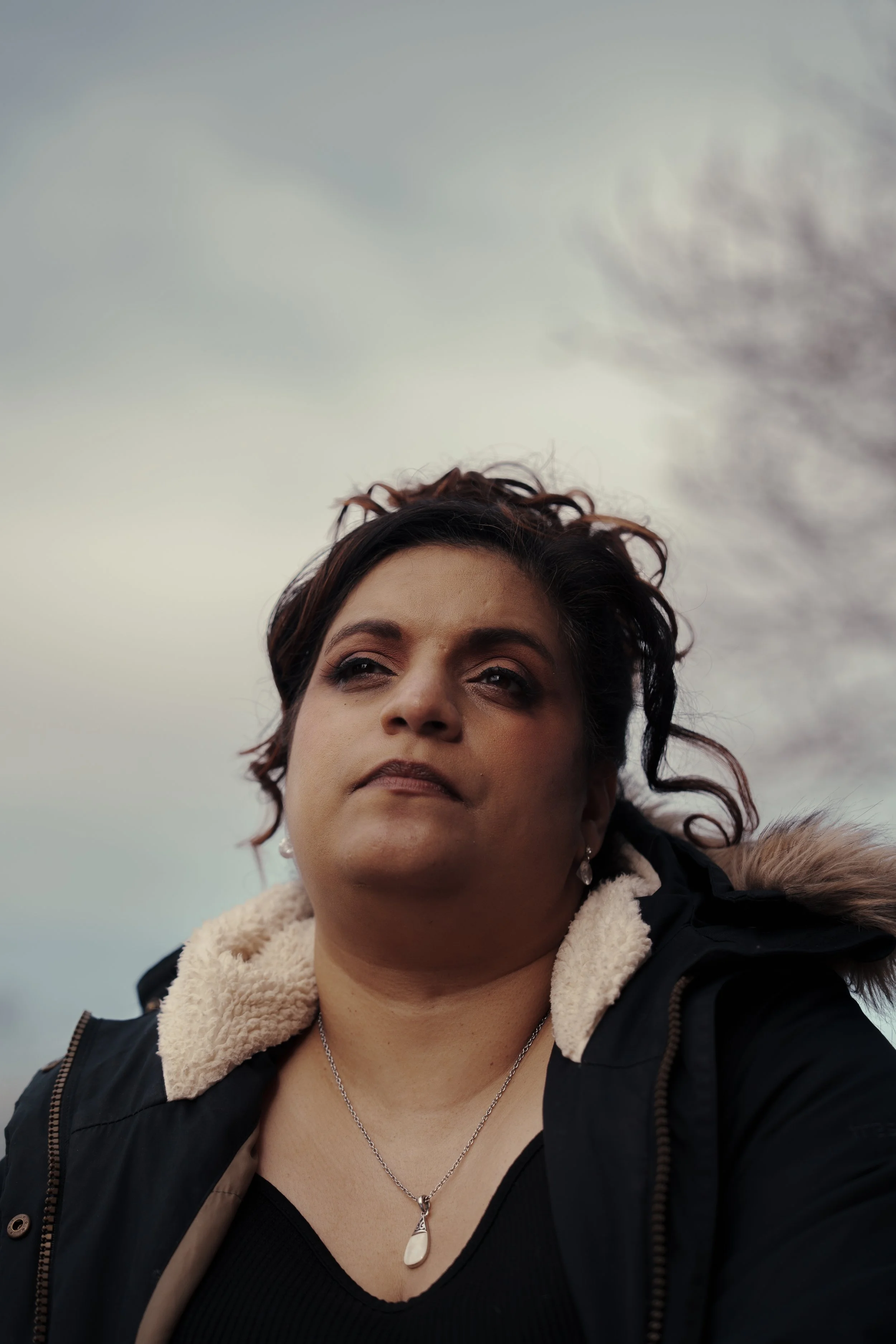 Close-up of a woman outdoors with cloudy sky in background, wearing a black jacket.