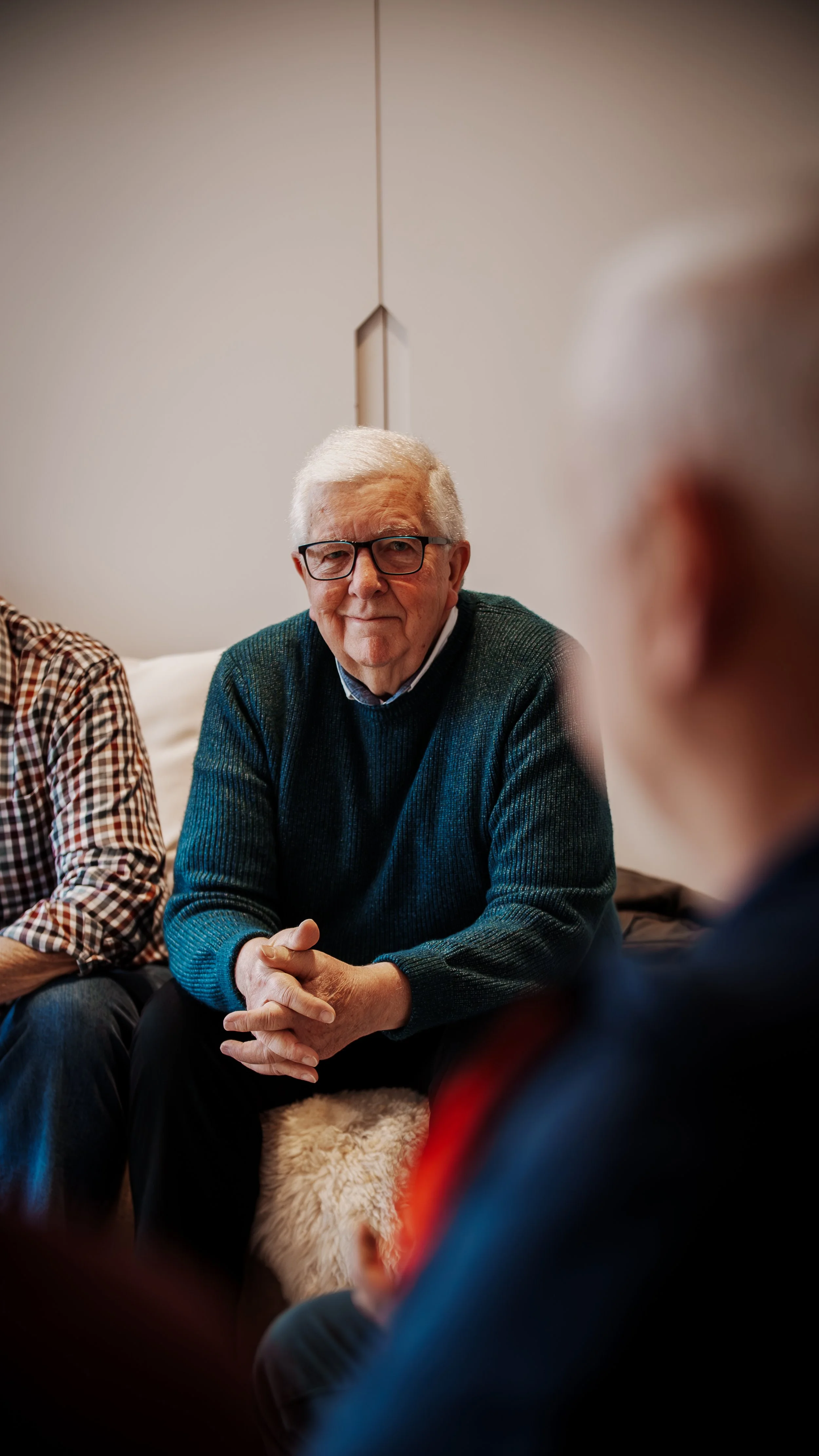 An elderly man with white hair and glasses, sitting on a chair with his hands clasped, in a cozy room, participating in a conversation with other people.