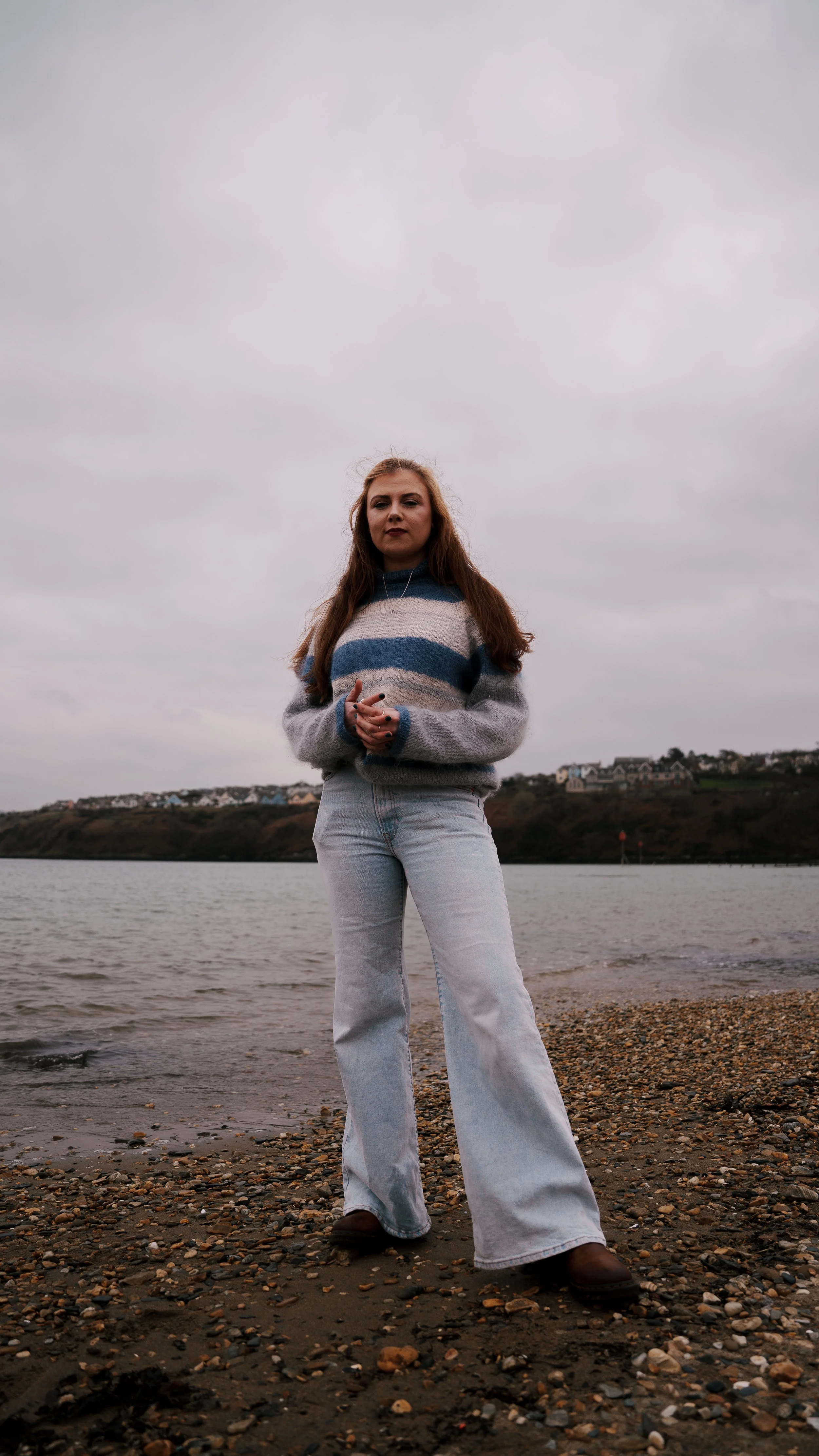 A young woman with long red hair stands on a pebbled beach near the sea under a cloudy sky, wearing a striped sweater and light-colored jeans.