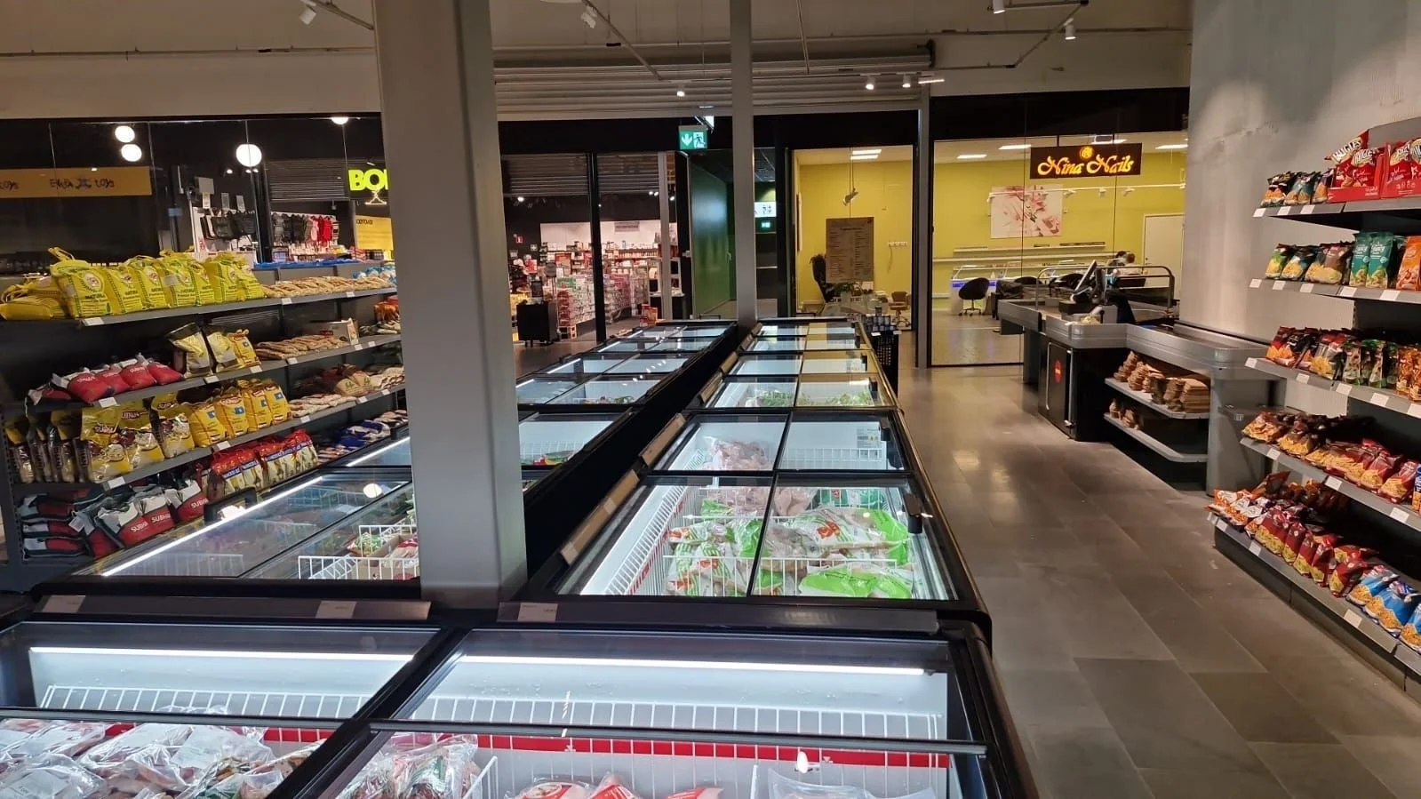 Inne i en mataffär med frysdiskar fulla av is och olika frysta varor, hyllor med snacks, och ett kassabord i bakgrunden.