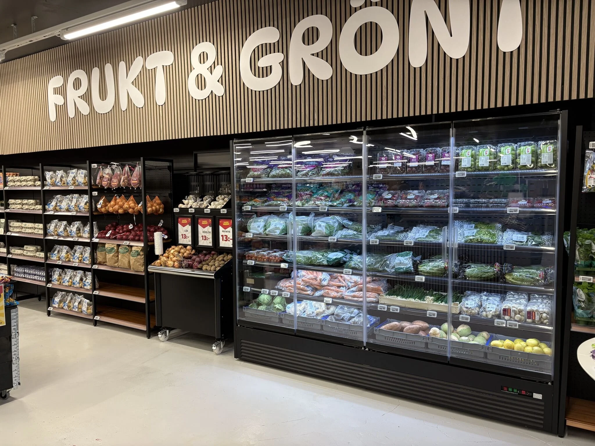 Frukt- och grönsaksavdelning i en mataffär med förvaringskyl och hyllor med olika grönsaker och frukt.