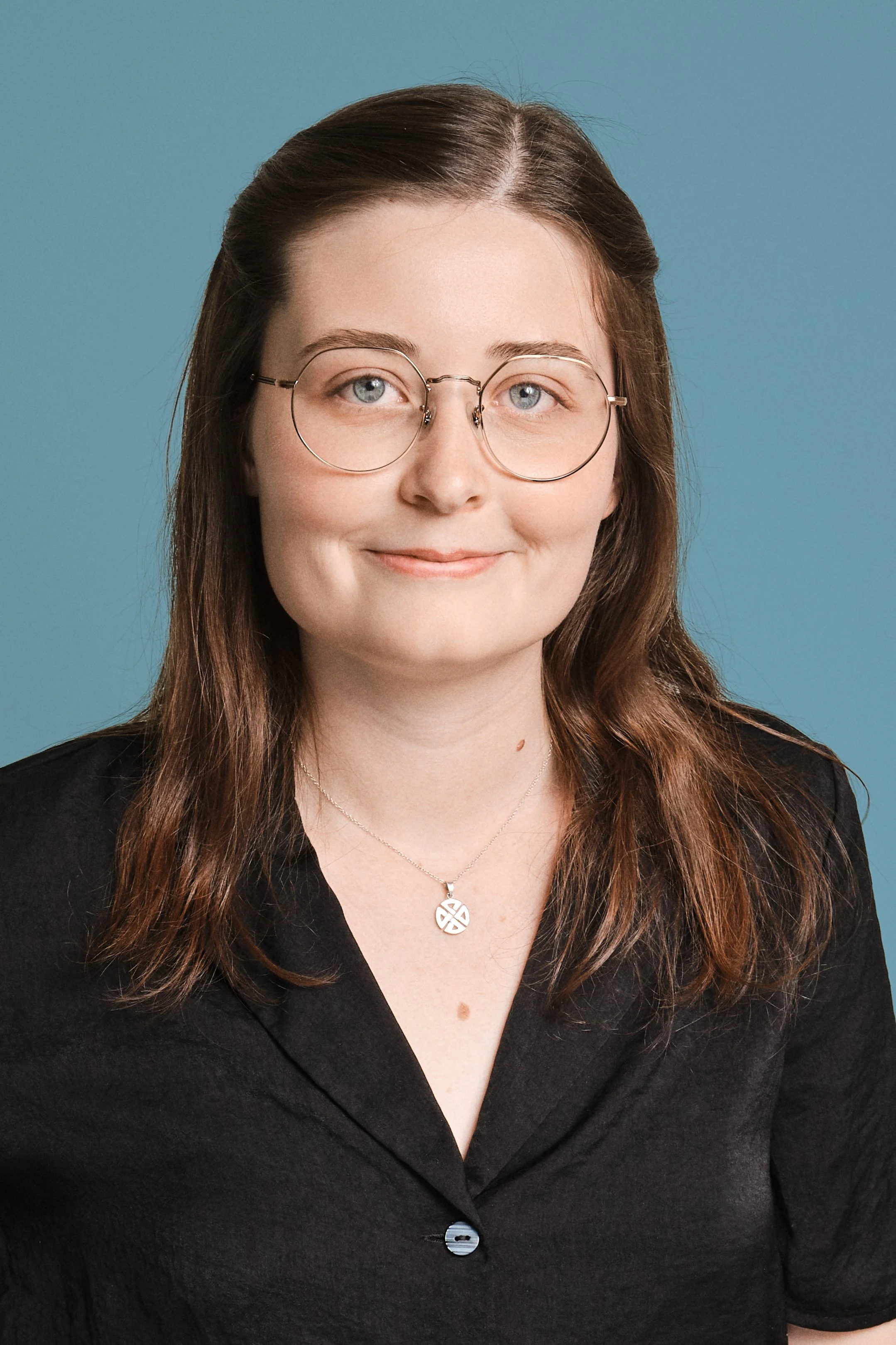 A young woman with brown hair, glasses, and a black blazer, smiling against a teal background.