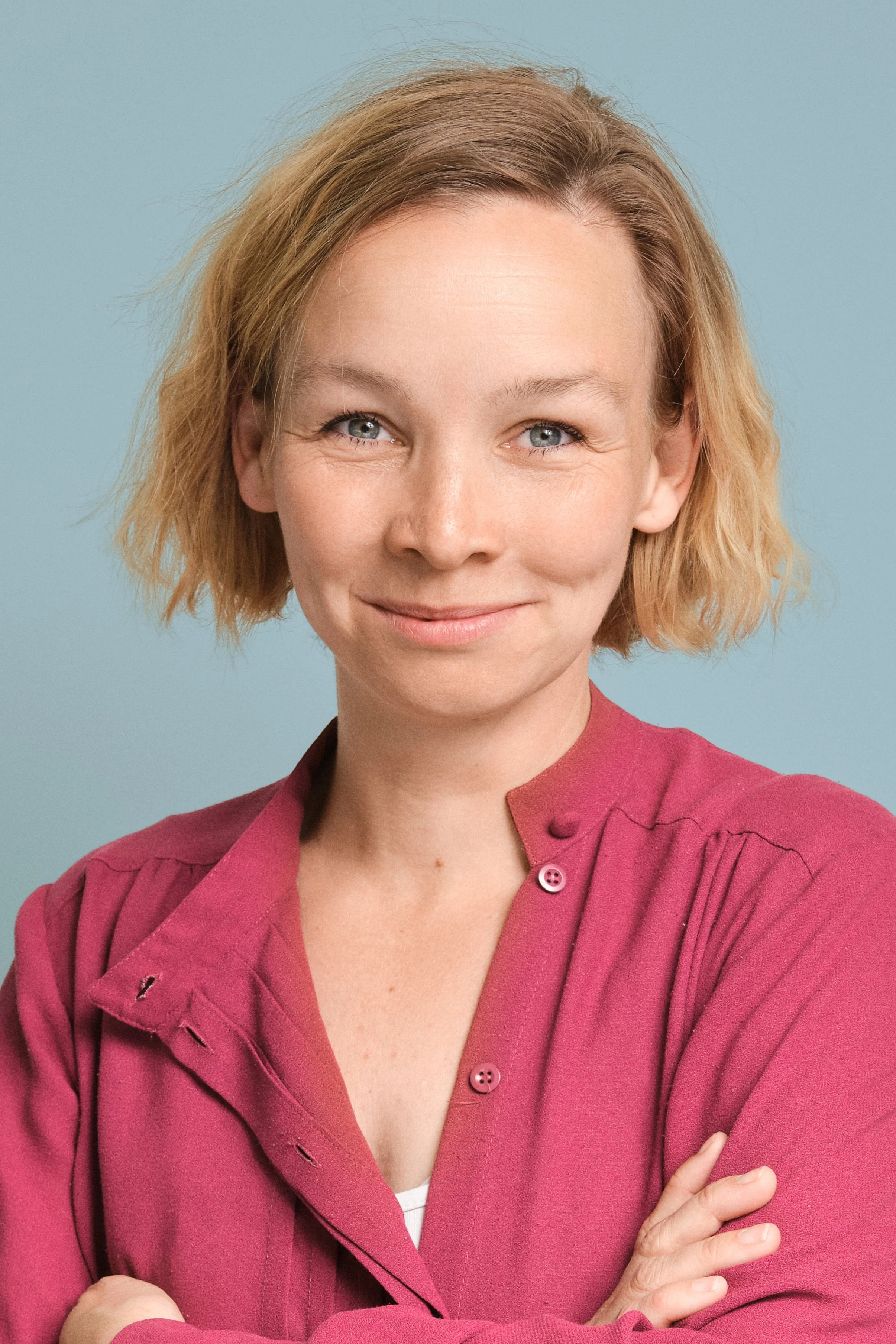 A woman with short blonde hair, blue eyes, and fair skin, wearing a pink button-up shirt with some holes in the shoulder, stands with her arms crossed against a light blue background.
