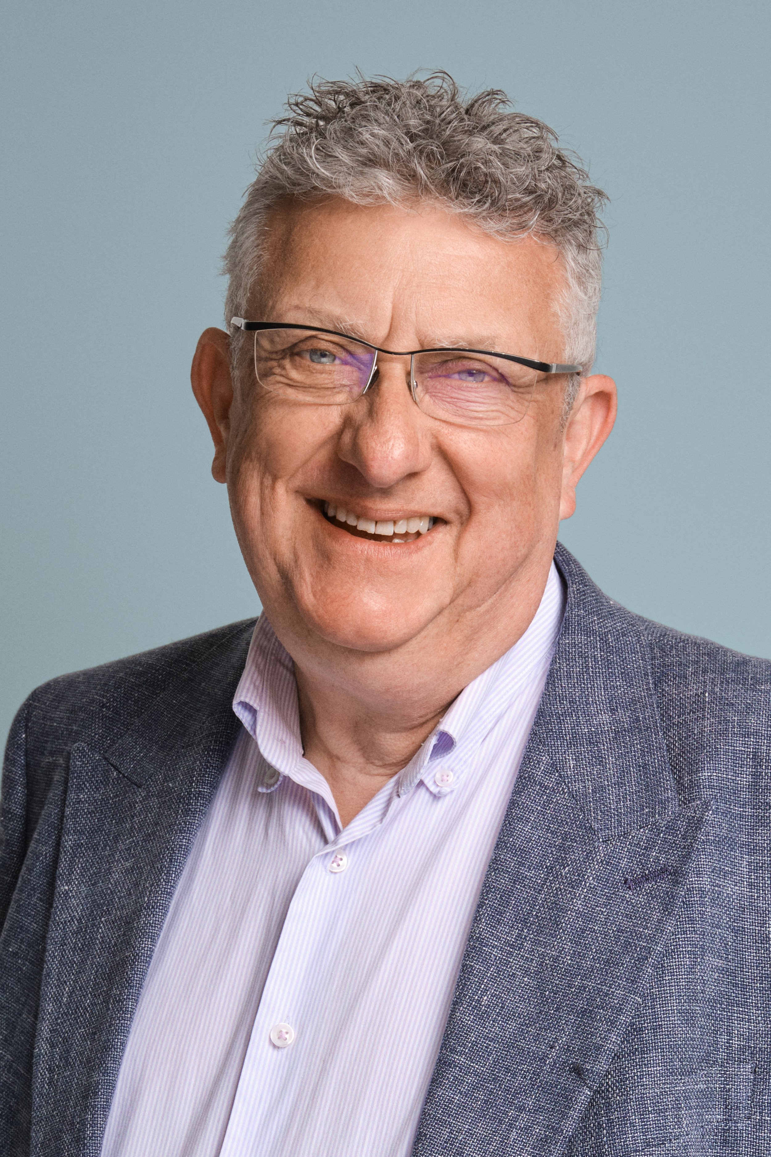 Headshot of a smiling middle-aged man with curly gray hair, glasses, and a gray blazer over a white shirt, against a plain light blue background.