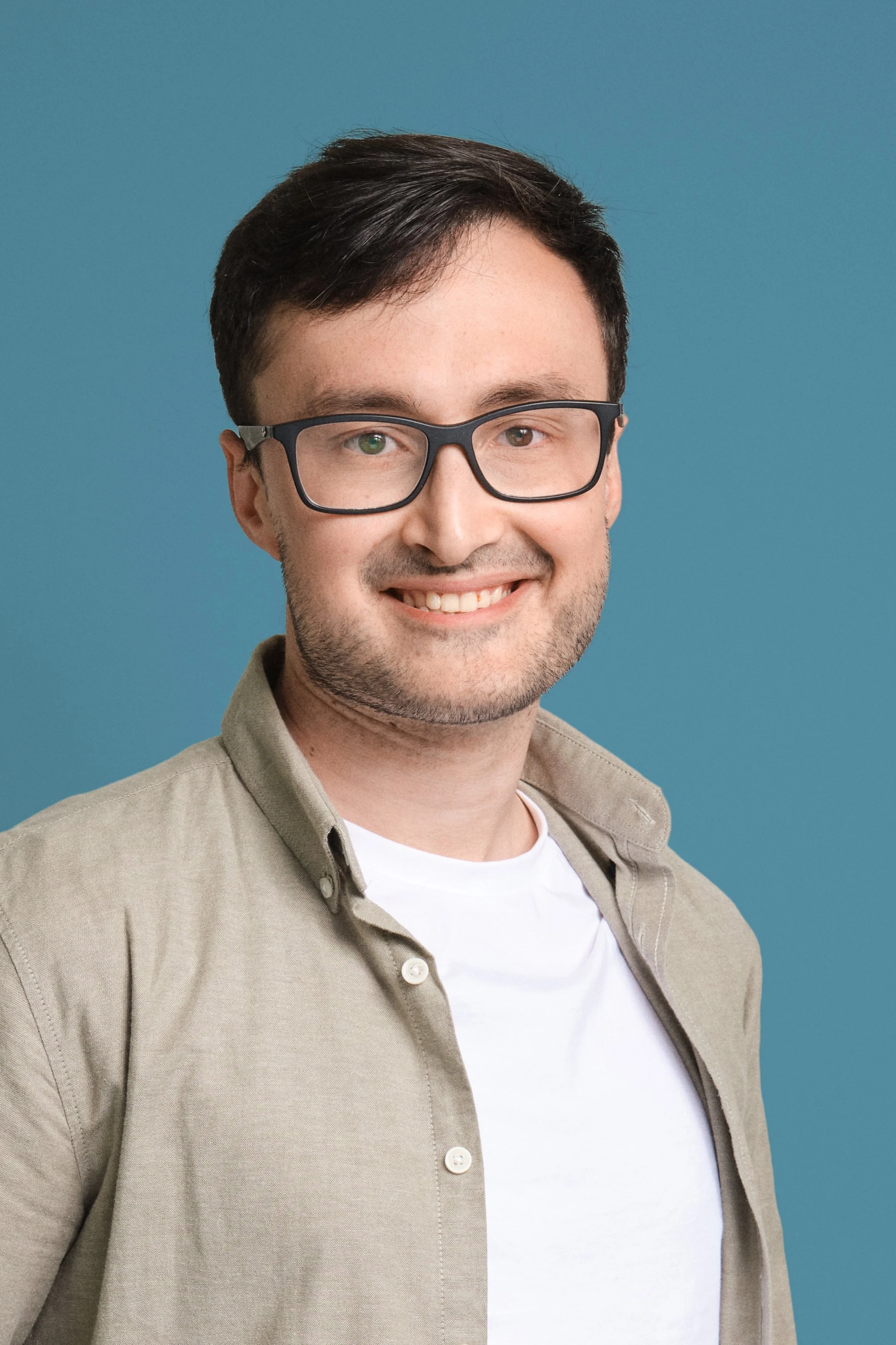 Portrait of a young man with glasses, smiling, wearing a beige shirt over a white t-shirt, against a blue background.