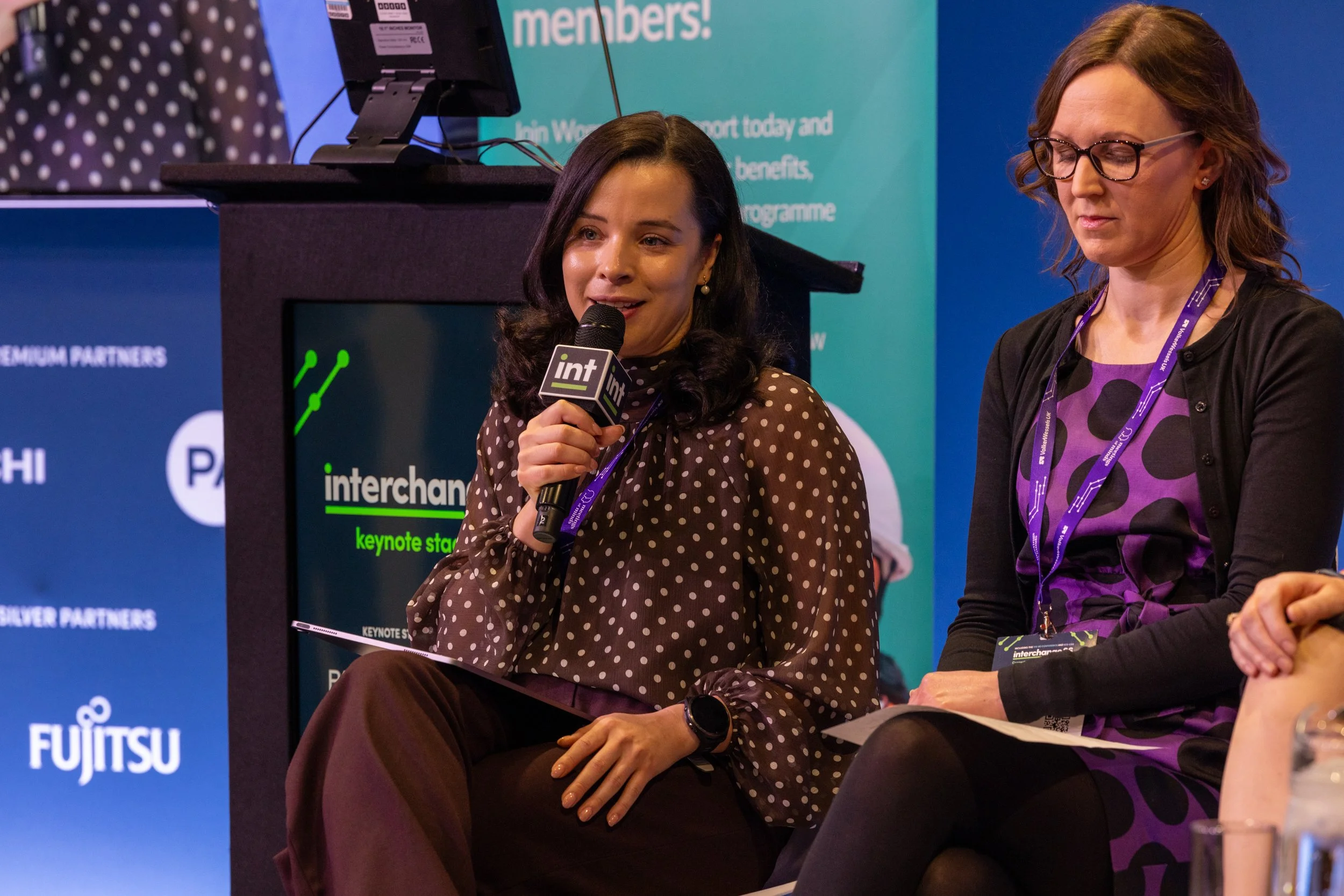 Two women sitting on a stage at a conference, one holding a microphone, with a blue and teal background with text and logos, and a monitor displaying the words "interchange keynote speaker".