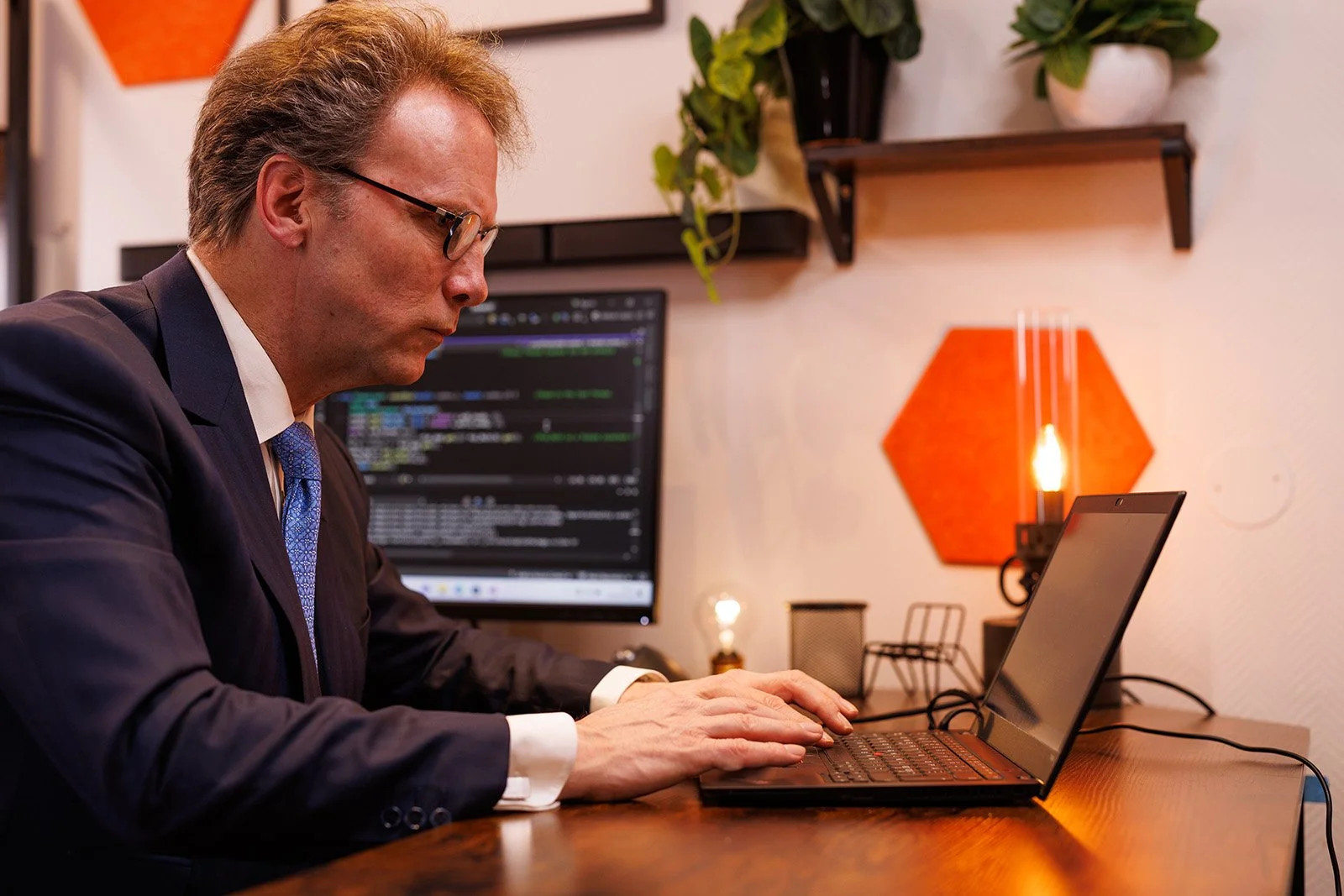 Man in a suit working on a laptop at a desk with a monitor displaying code, plants, and decorative wall items.