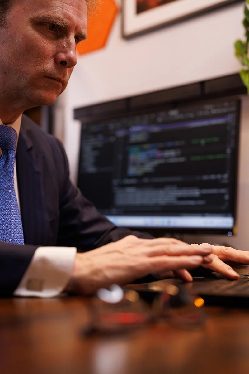Person in a suit typing on a computer keyboard, with code visible on the monitor in the background.