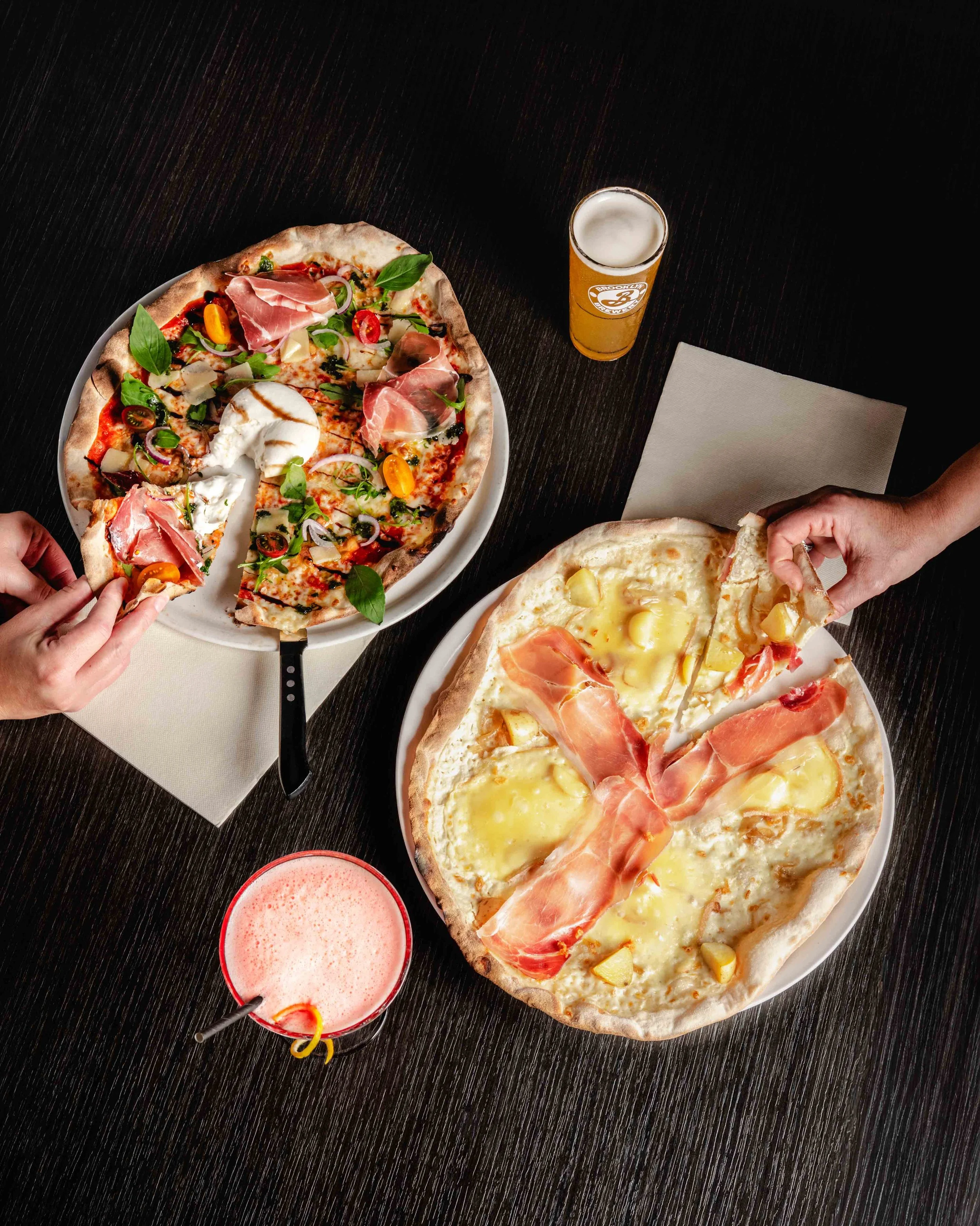 Deux pizzas sur des assiettes, des boissons et deux mains en train de prendre une part de pizza. Issue d'un projet de photo culinaire du Studio Kroma pour le restaurant L'Eden Bowl.