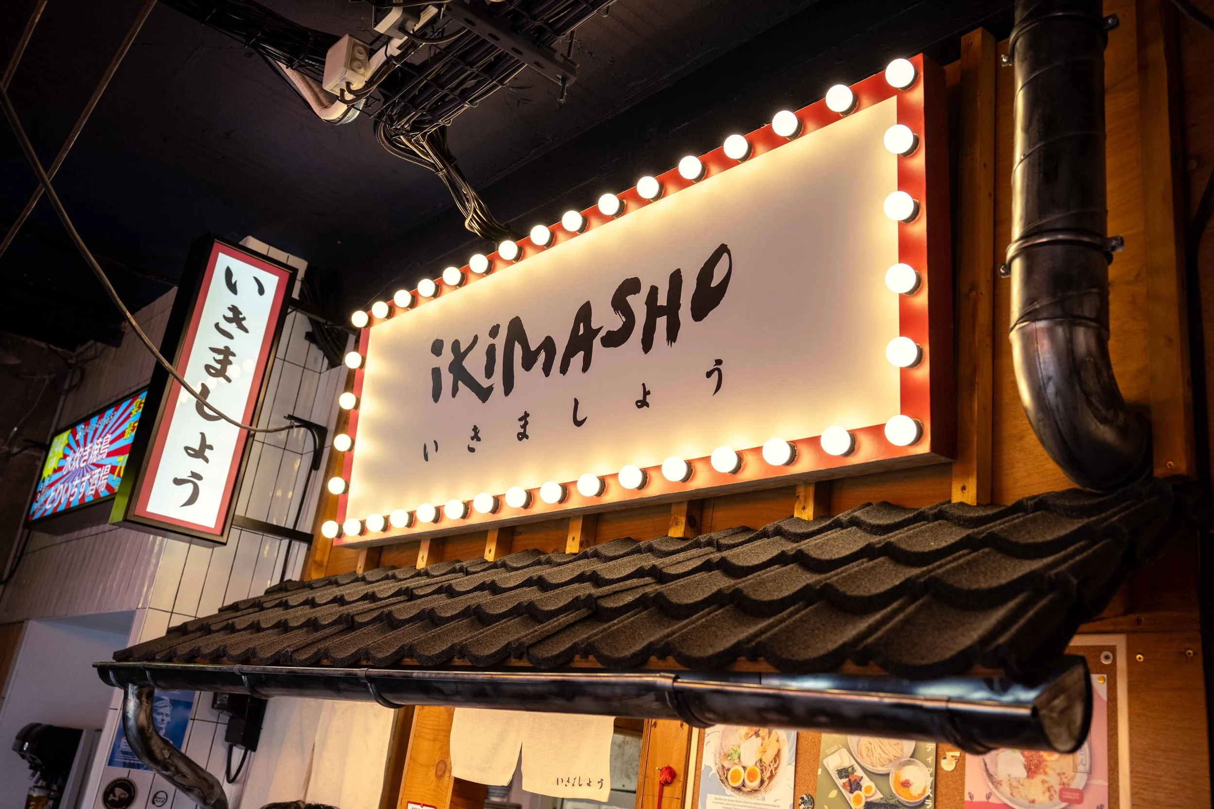 Un panneau lumineux avec le nom 'IKIMASHO' écrit en anglais et japonais, situé à l'extérieur d'un restaurant japonais, décoré de lumières blanches. Issue du spot publicitaire du Studio Kroma pour le restaurant Ikimasho.