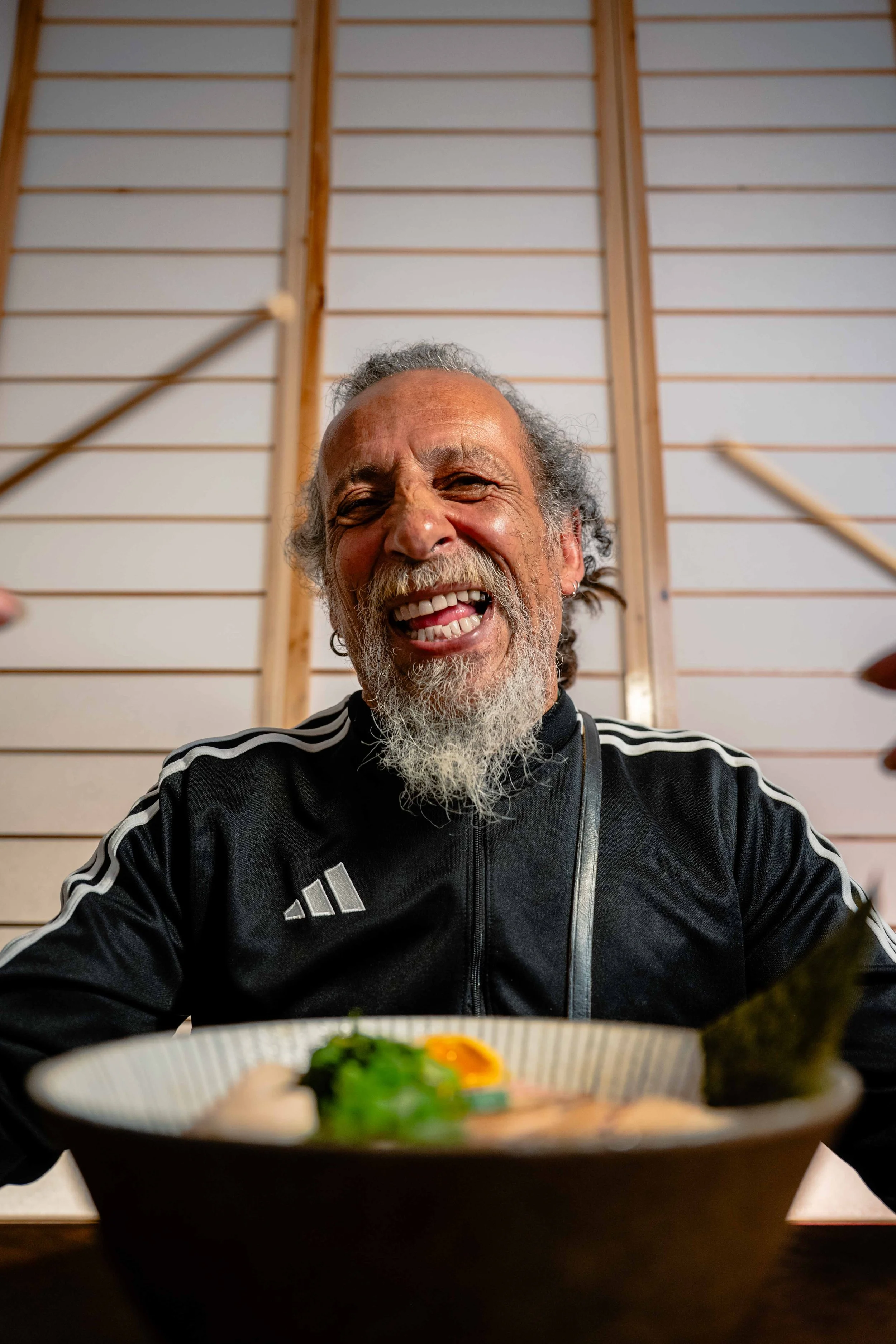 Homme âgé souriant avec une barbe blanche, portant un maillot noir avec des bandes blanches, assis devant un bol de ramen dans un environnement intérieur en bois. Issue du spot publicitaire du Studio Kroma pour le restaurant Ikimasho.