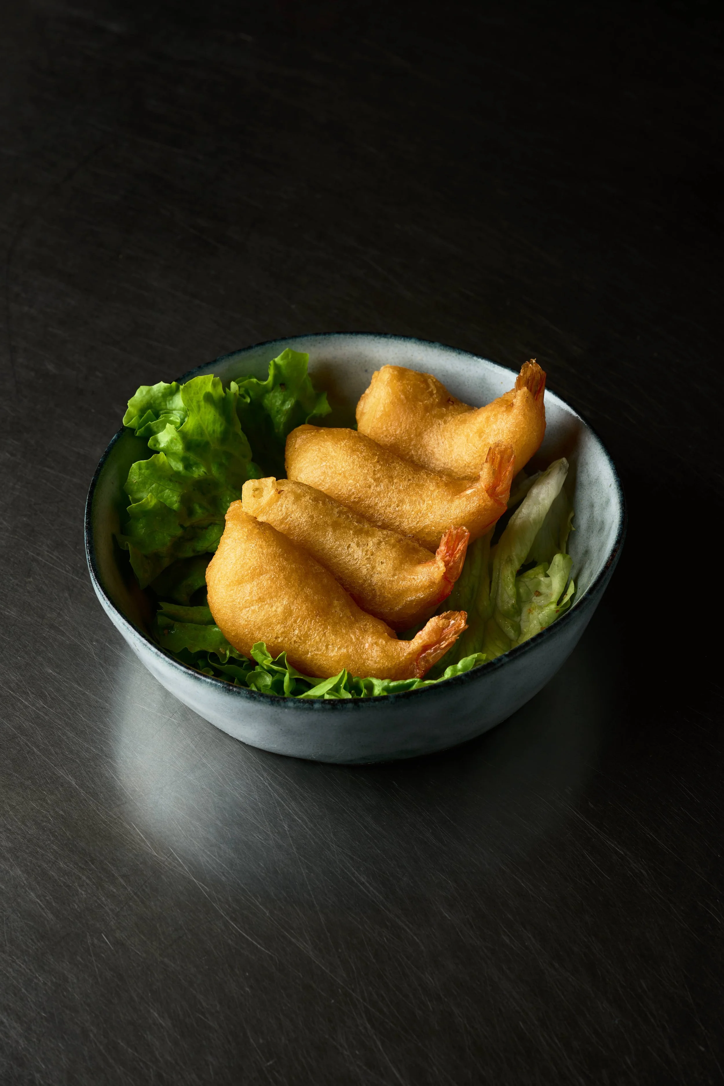 Un bol noir et blanc contenant quatre crevettes frites sur de la laitue verte, posé sur une table sombre. Issue d'un projet de photo culinaire du Studio Kroma pour le traiteur HA MY de Geneston.