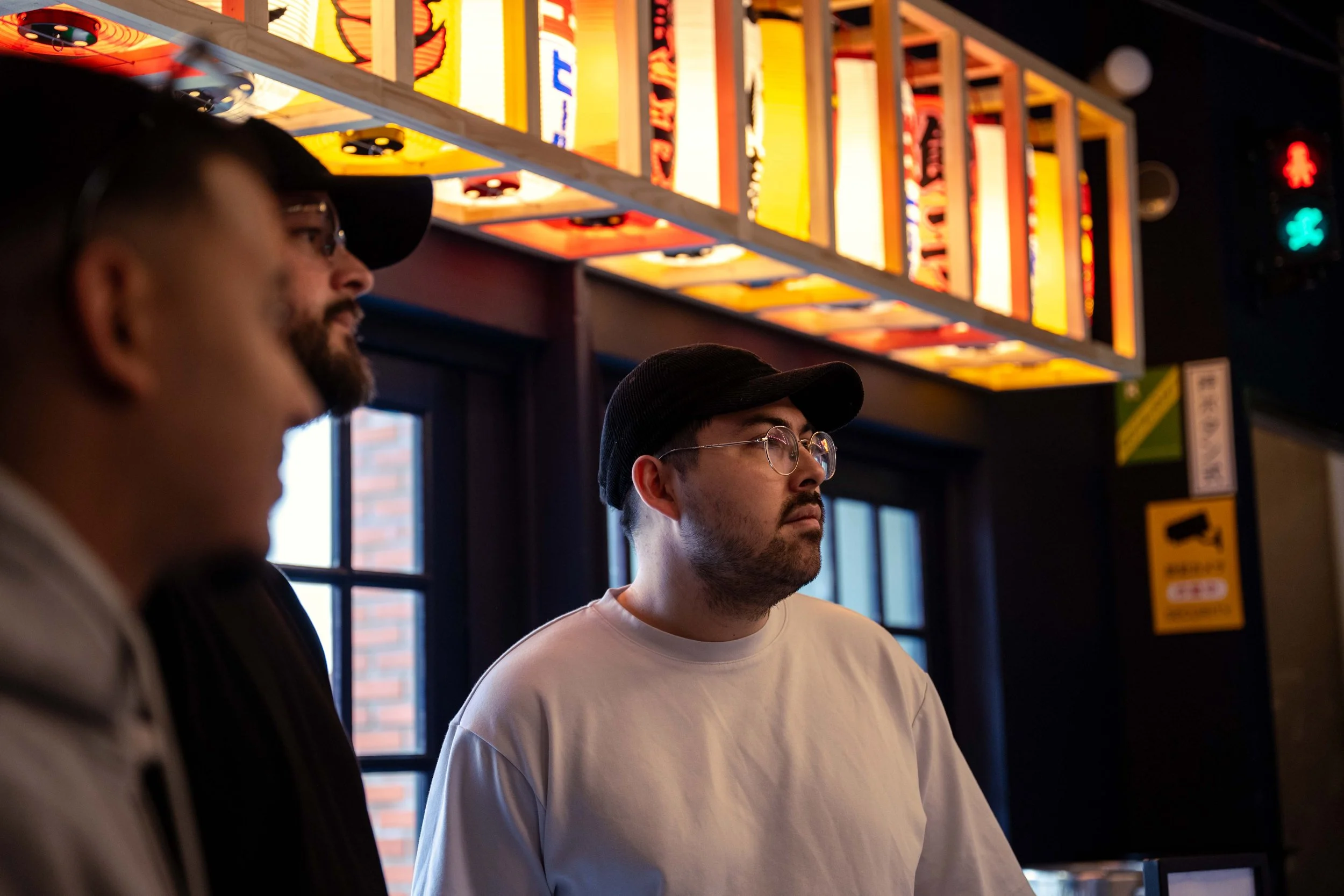 Trois hommes dans un restaurant ou bar, un homme avec lunettes, casquette noire, et chemise blanche, en arrière-plan au-dessus, enseigne lumineuse colorée.