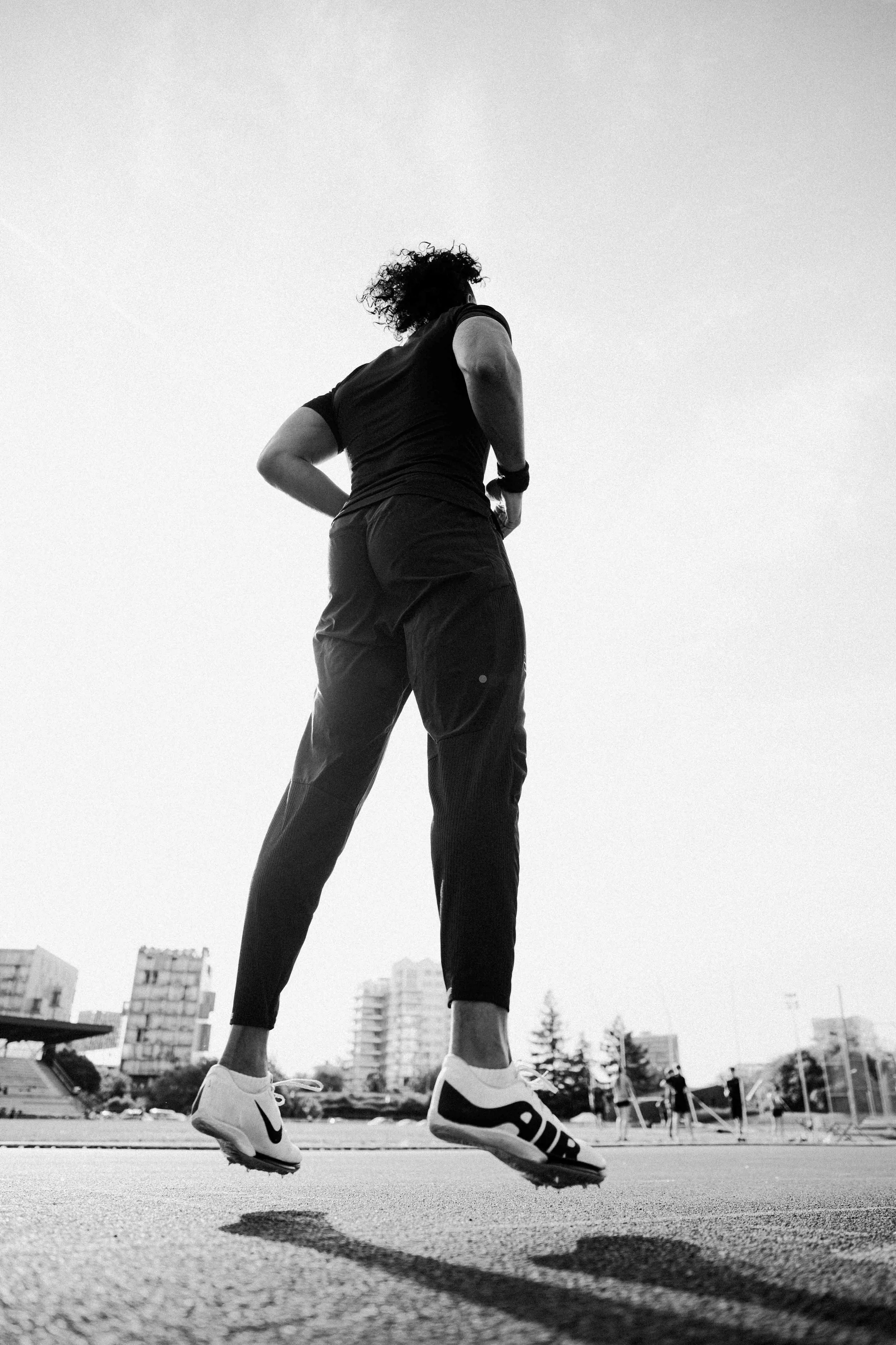 Une femme, vue de dos, en train de sauter sur une piste d'athlétisme en extérieur, en noir et blanc, avec un paysage urbain en arrière-plan. Issue d'une vidéo de sport du Studio Kroma pour le projet Limitless.