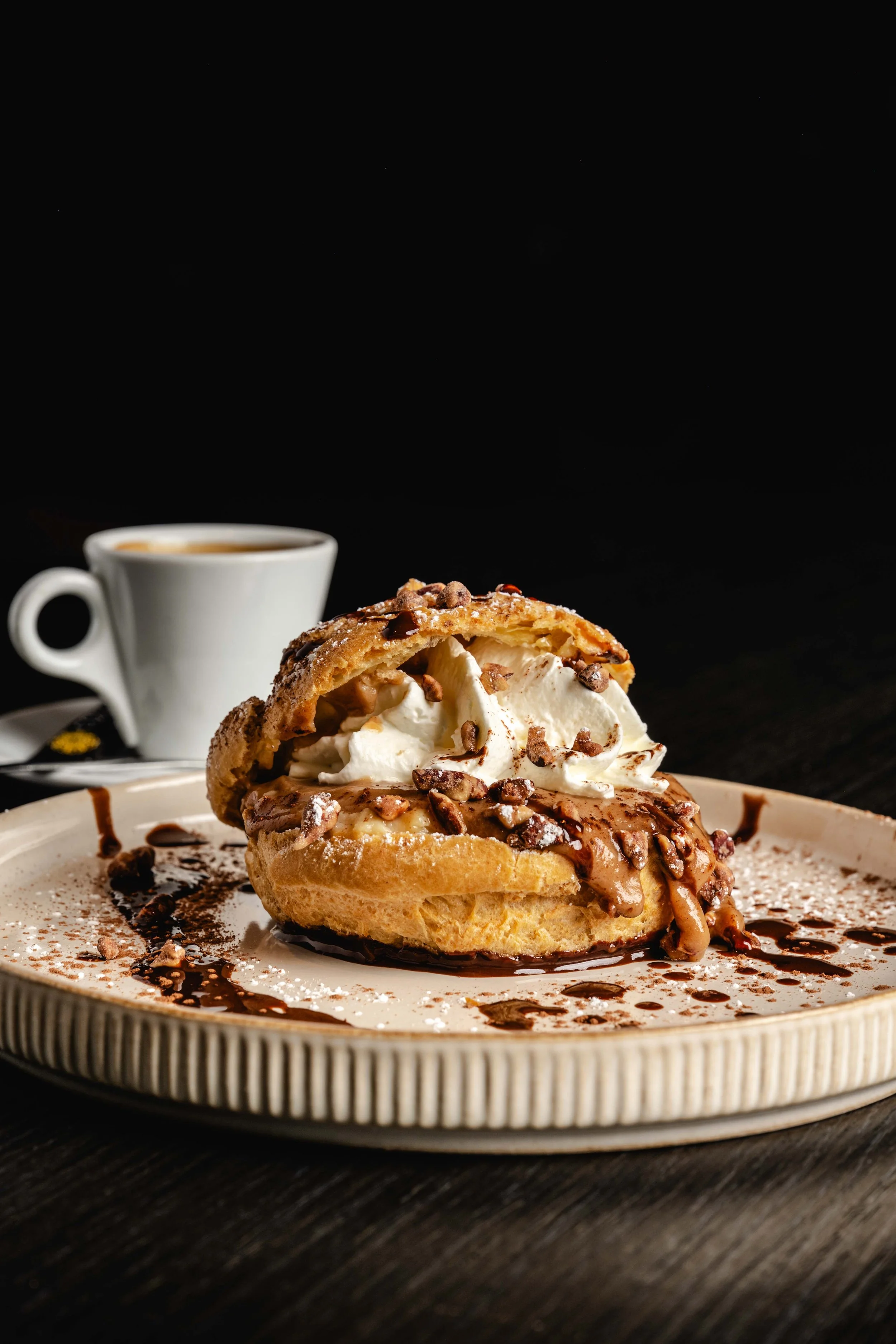 Chou fourrée à la crème fouettée, chocolat et noisettes, servie sur une assiette avec du chocolat et une tasse de café en arrière-plan.  Issue d'un projet de photo culinaire du Studio Kroma pour le restaurant L'Eden Bowl.