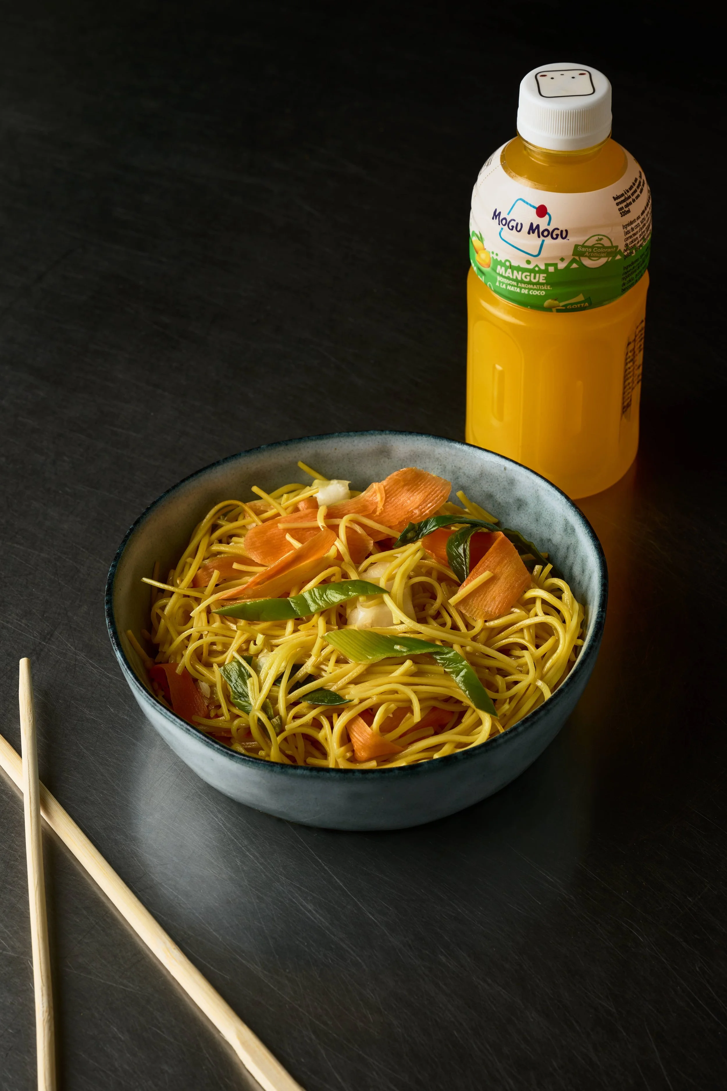 Bol de noodles avec légumes verts et carottes, bouteille de jus de mangue, et une paire de baguettes sur une table noire. Issue d'un projet de photo culinaire du Studio Kroma pour le traiteur HA MY de Geneston.