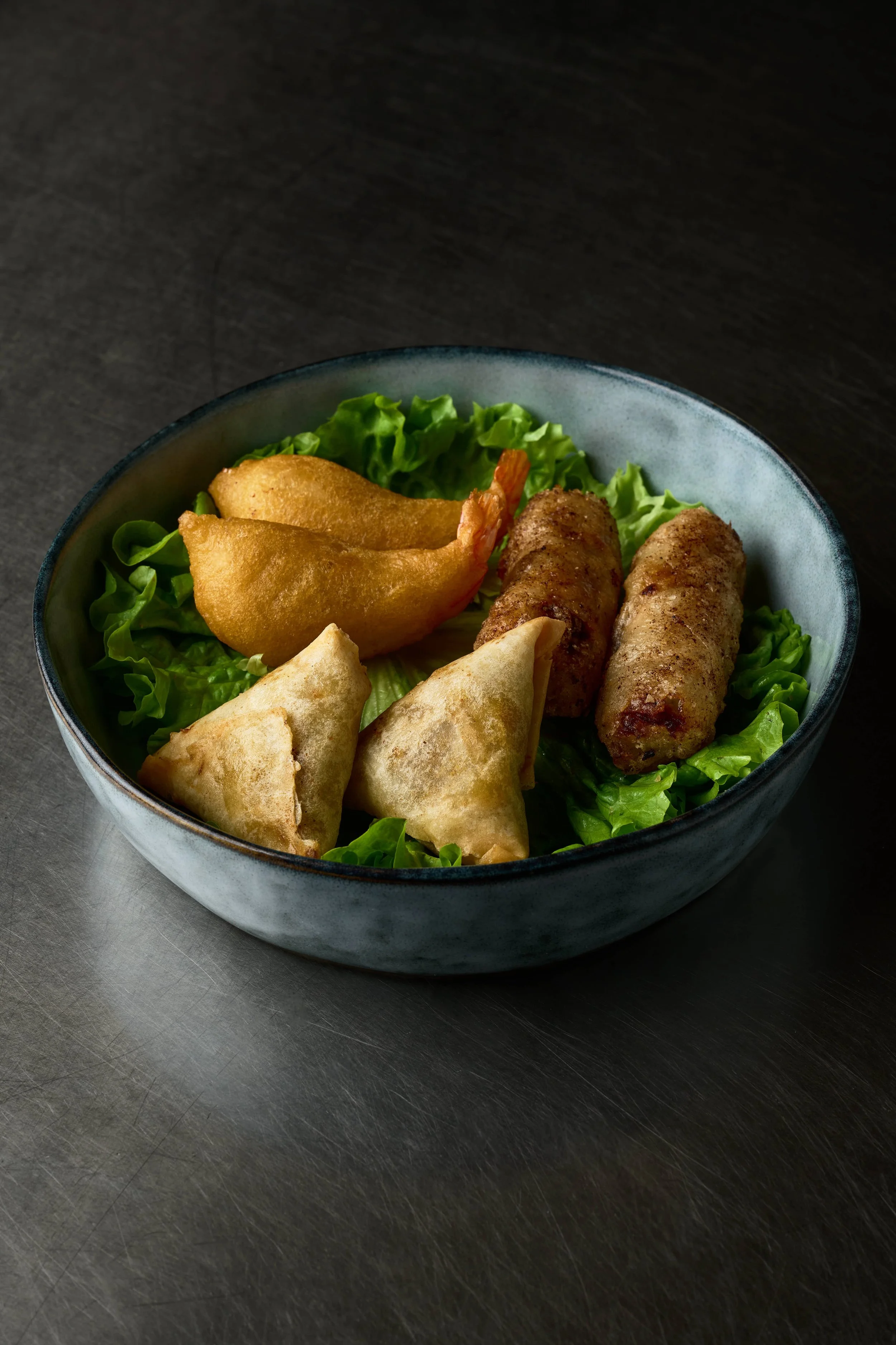 Un bol contenant des nems, des bâtonnets de poulet frit, des triangles de pâte frit, et des morceaux de crevettes frites, servi sur un lit de laitue. Issue d'un projet de photo culinaire du Studio Kroma pour le traiteur HA MY de Geneston.