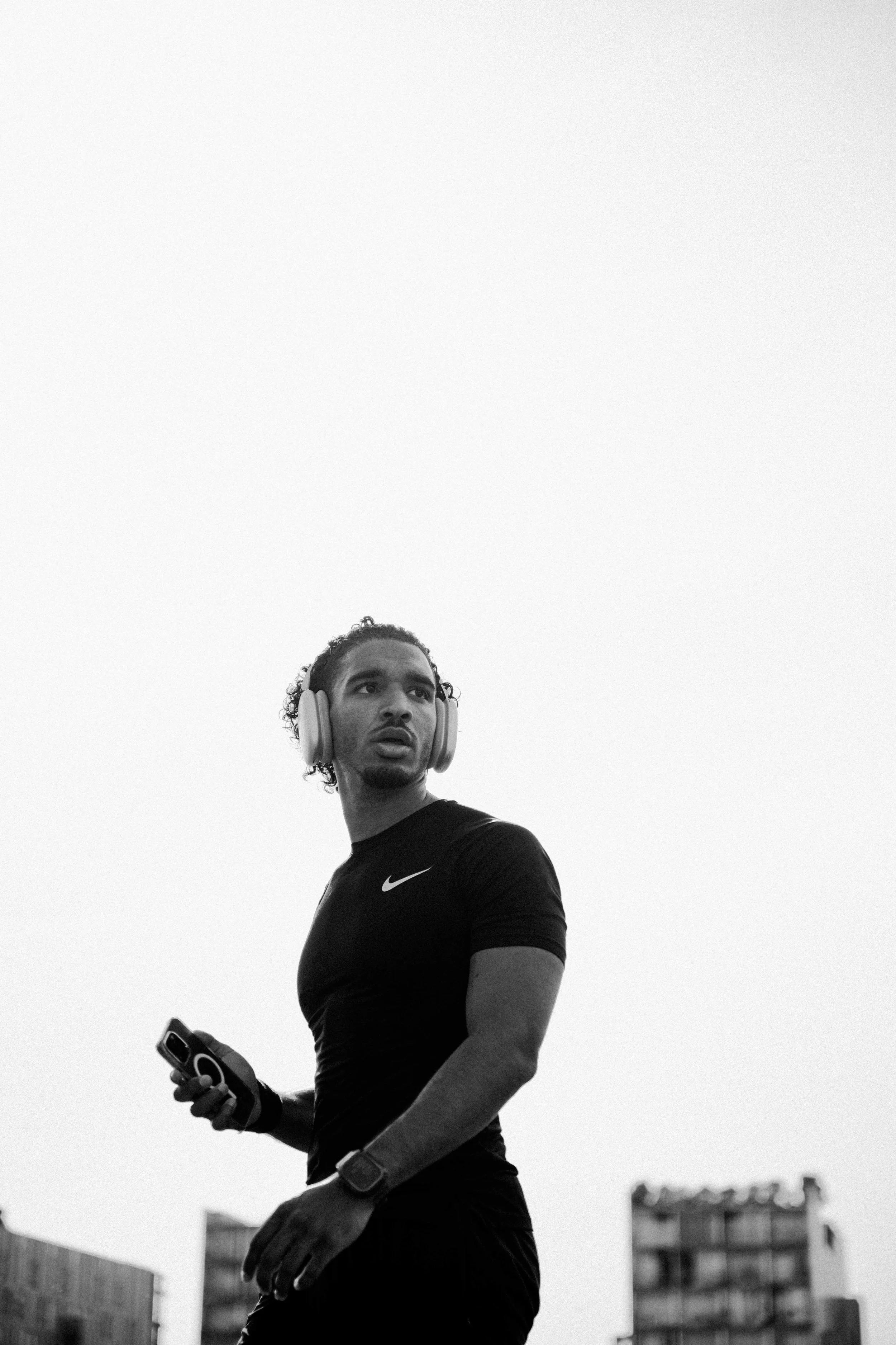 Un homme portant un t-shirt noir avec un logo Nike, portant des écouteurs et une montre, tenant un appareil électronique, avec un fond de bâtiments urbains.