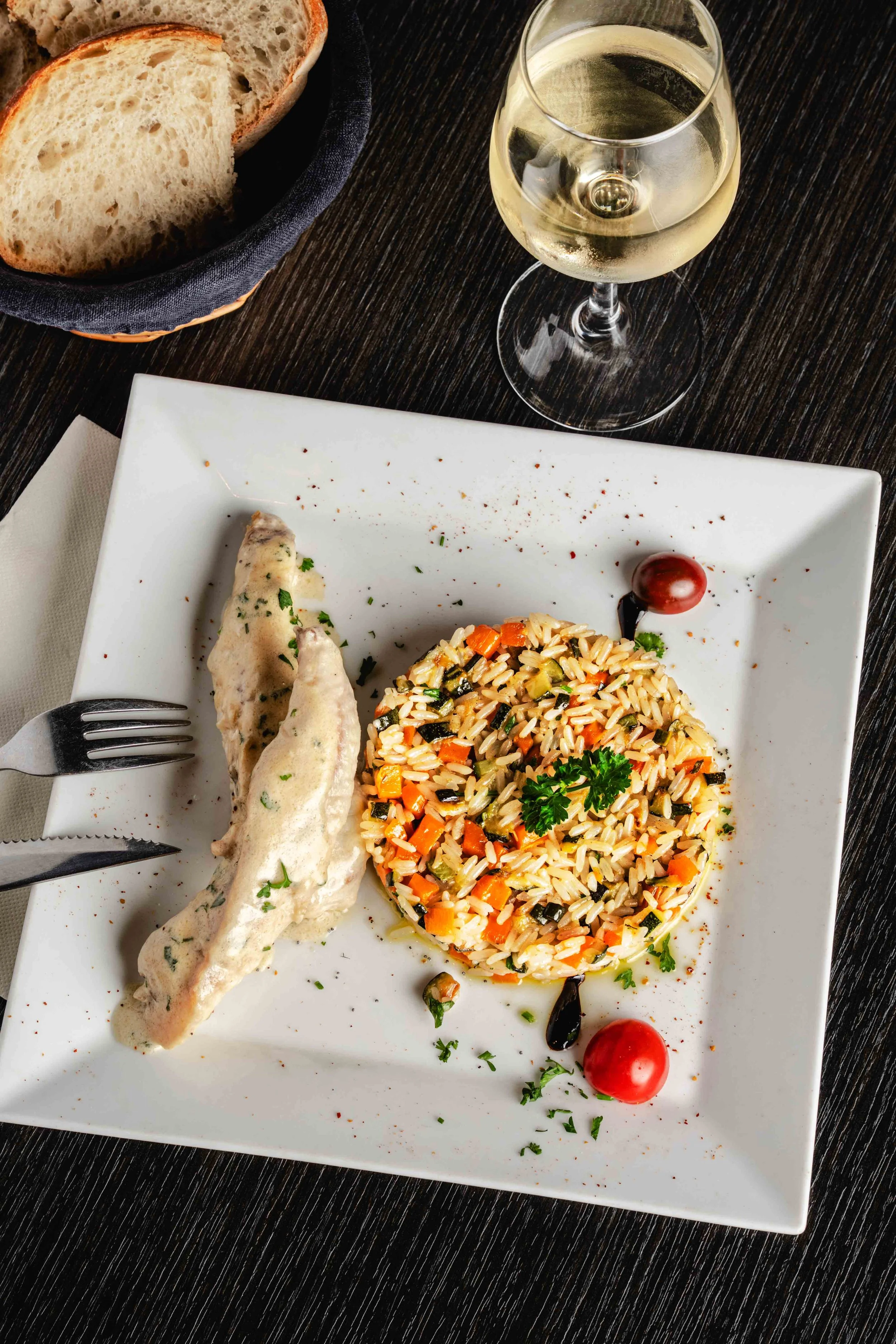 Assiette de riz aux légumes avec un morceau de poisson en sauce, accompagnée d'un verre de vin blanc, pain en tranches et tomates cerises, sur une table en bois.  Issue d'un projet de photo culinaire du Studio Kroma pour le restaurant L'Eden Bowl.