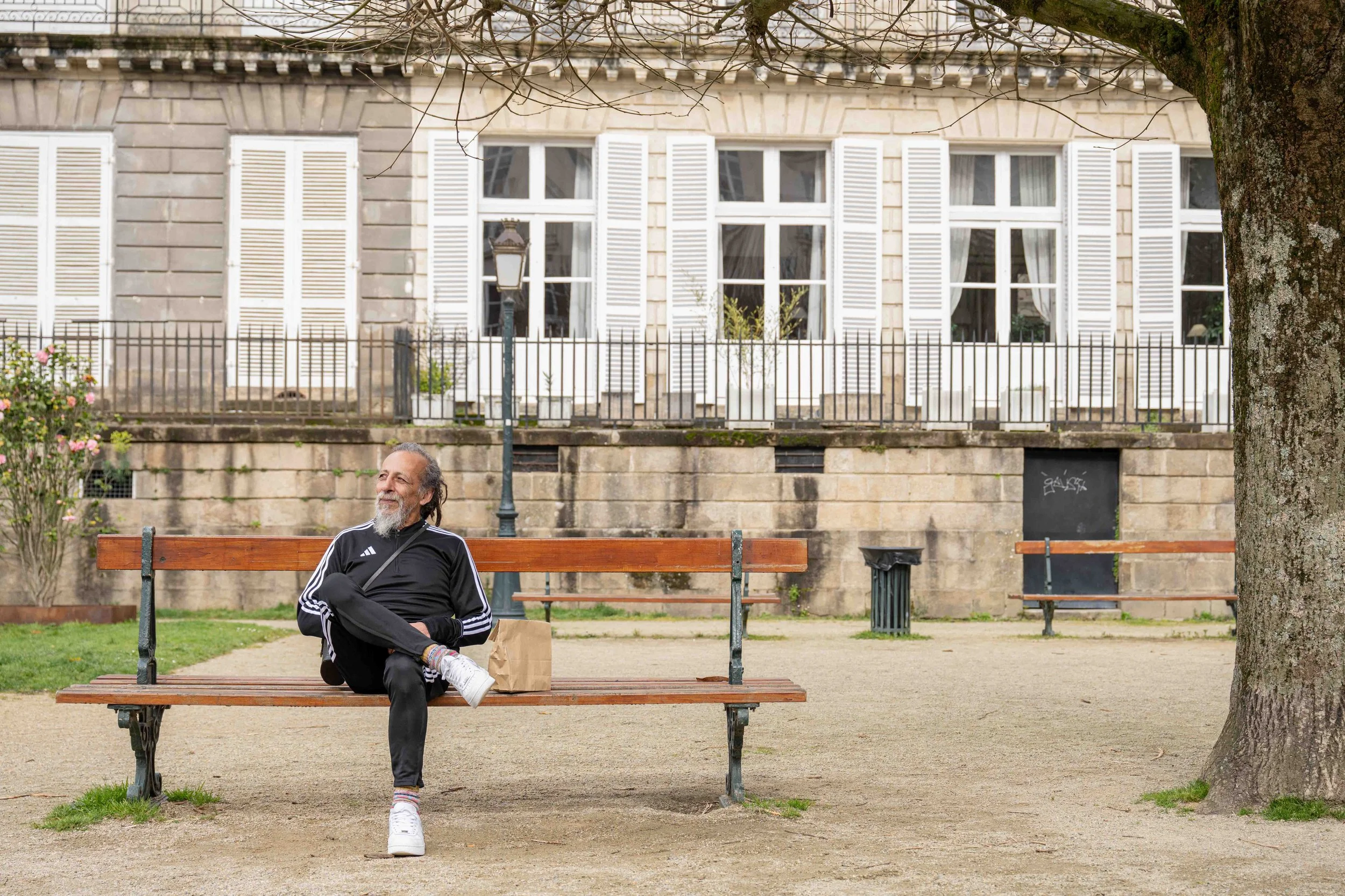 Un homme âgé assis sur un banc dans un parc, souriant, avec en arrière-plan une maison avec des fenêtres à volets blancs. Issue du spot publicitaire du Studio Kroma pour le restaurant Ikimasho.