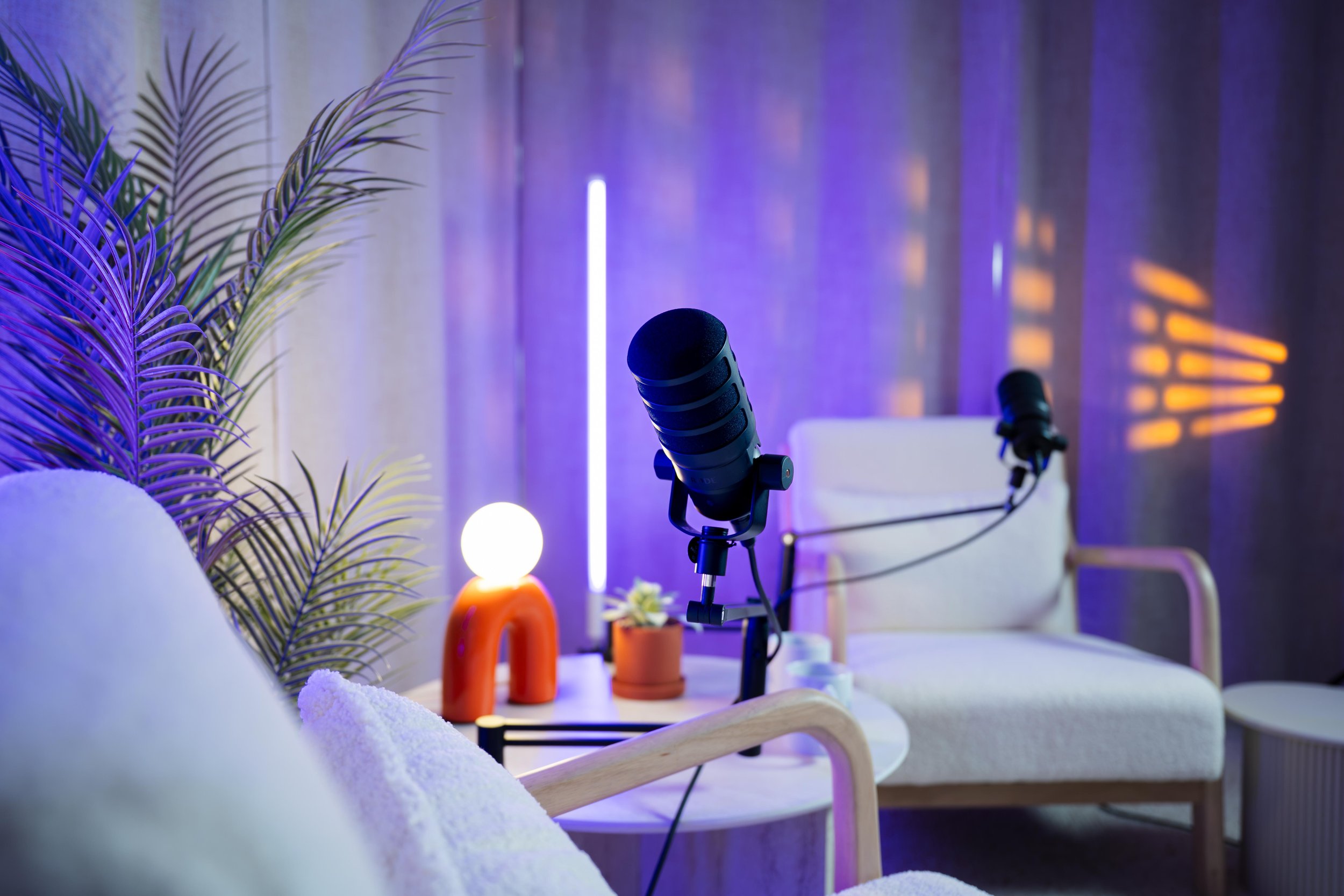 Photo du studio de podcasts et formats verticaux du Studio Kroma situé à Vertou. Deux microphones dans un studio d'enregistrement avec des fauteuils beige, des plantes et des lampes décoratives en arrière-plan, éclairage violet et orange.