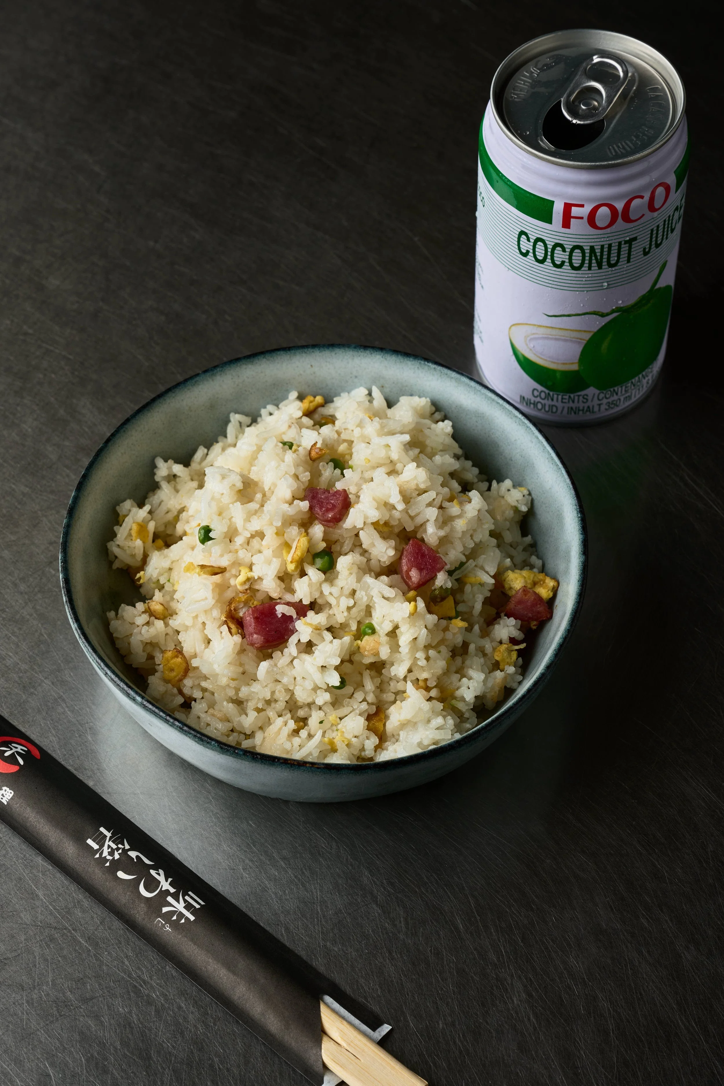 Bol de riz frit avec petits morceaux de viande et œufs, canette de jus de noix de coco, et paire de baguettes noires sur une surface sombre.  Issue d'un projet de photo culinaire du Studio Kroma pour le traiteur HA MY de Geneston.
