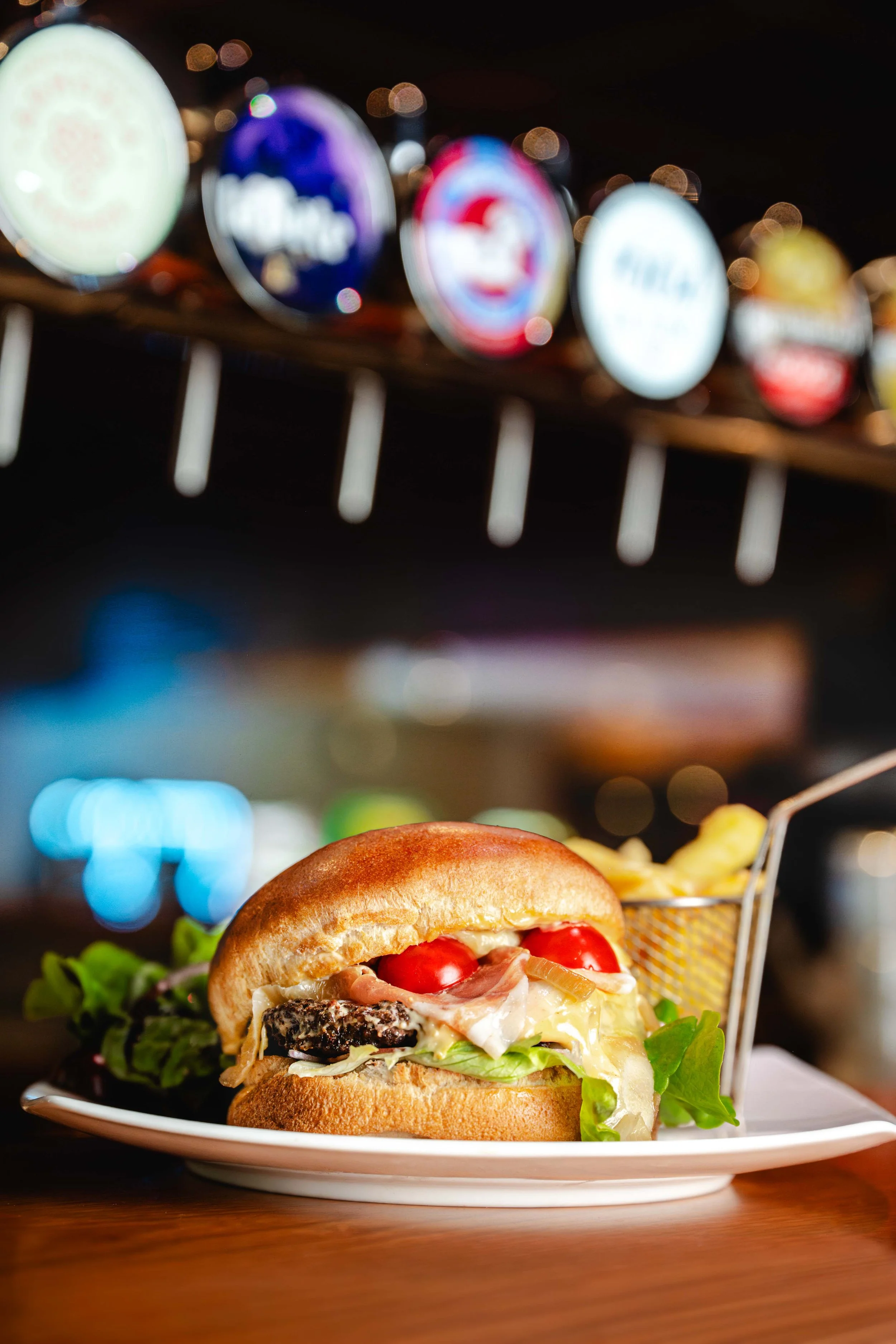 Sandwich avec pain brioche, tomates cerises, salade, fromage, jambon, et un côté de frites dans une petite cage en métal, sur une assiette blanche. Issue d'un projet de photo culinaire du Studio Kroma pour le restaurant L'Eden Bowl.