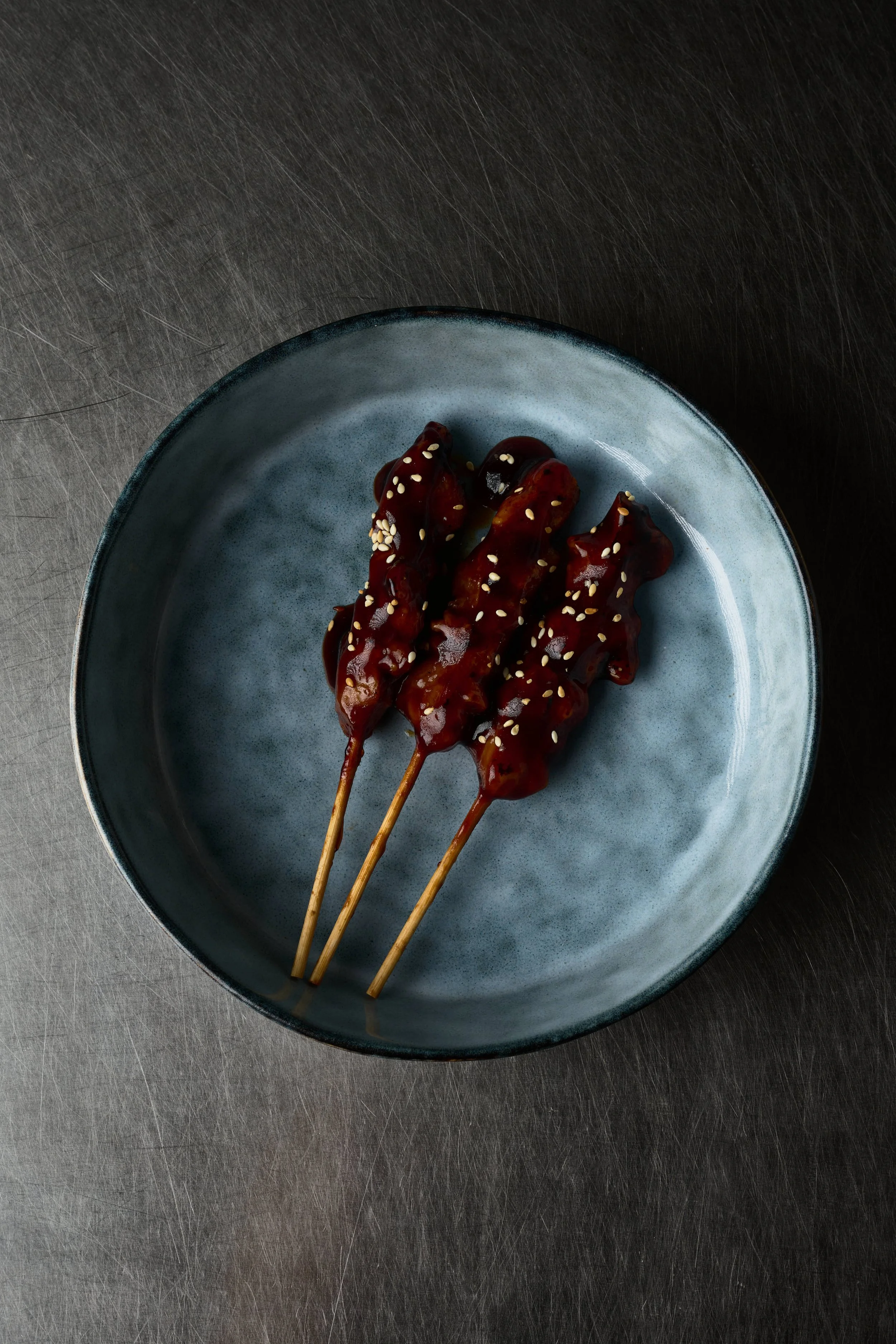 Trois brochettes de viande enrobées de sauce brune et saupoudrées de graines de sésame, servies dans une assiette bleue sur une surface en acier. Issue d'un projet de photo culinaire du Studio Kroma pour le traiteur HA MY de Geneston.