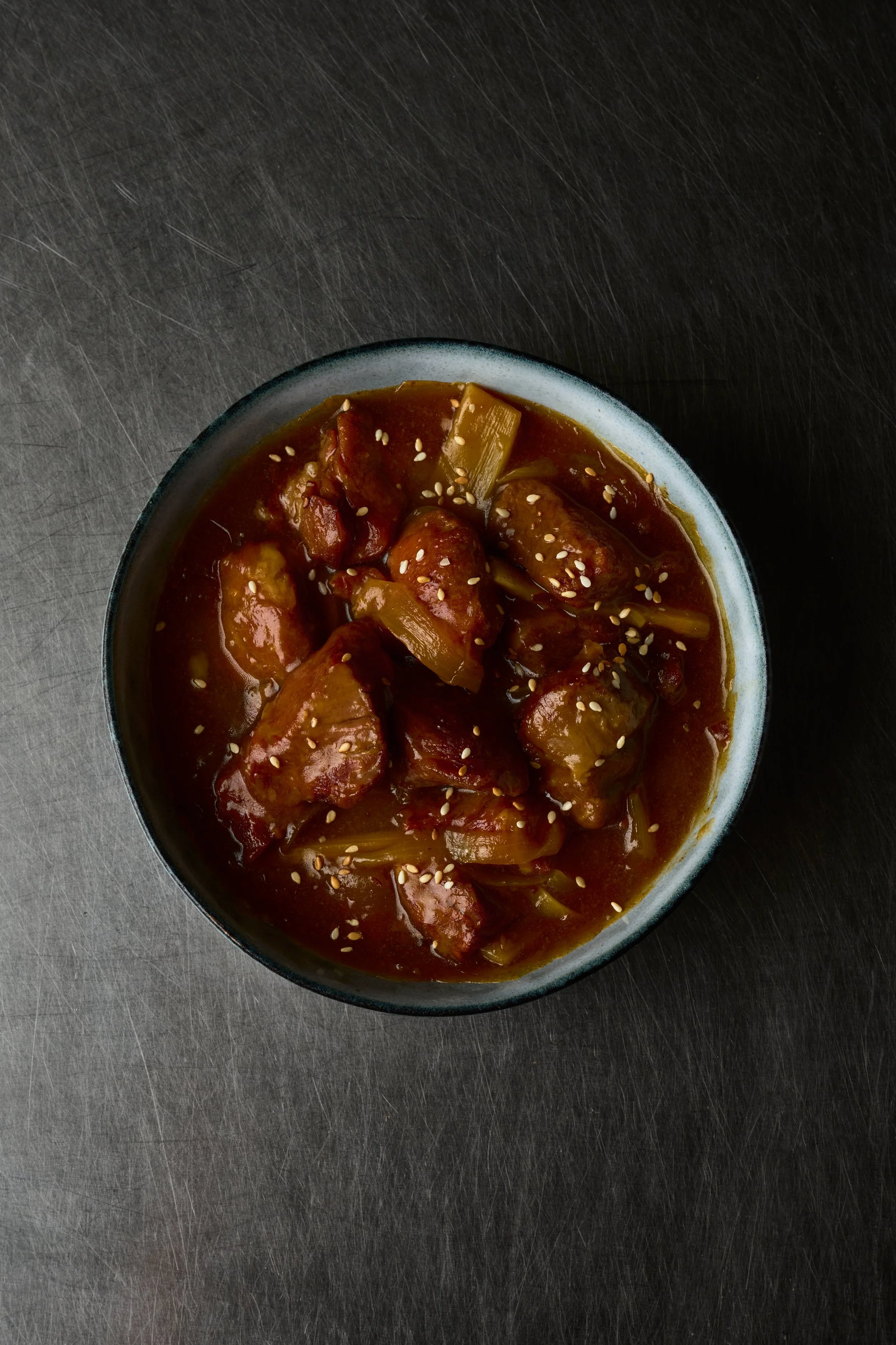 Bol de viande et légumes en sauce, garnie de graines de sésame, sur une surface sombre en métal. Issue d'un projet de photo culinaire du Studio Kroma pour le traiteur HA MY de Geneston.