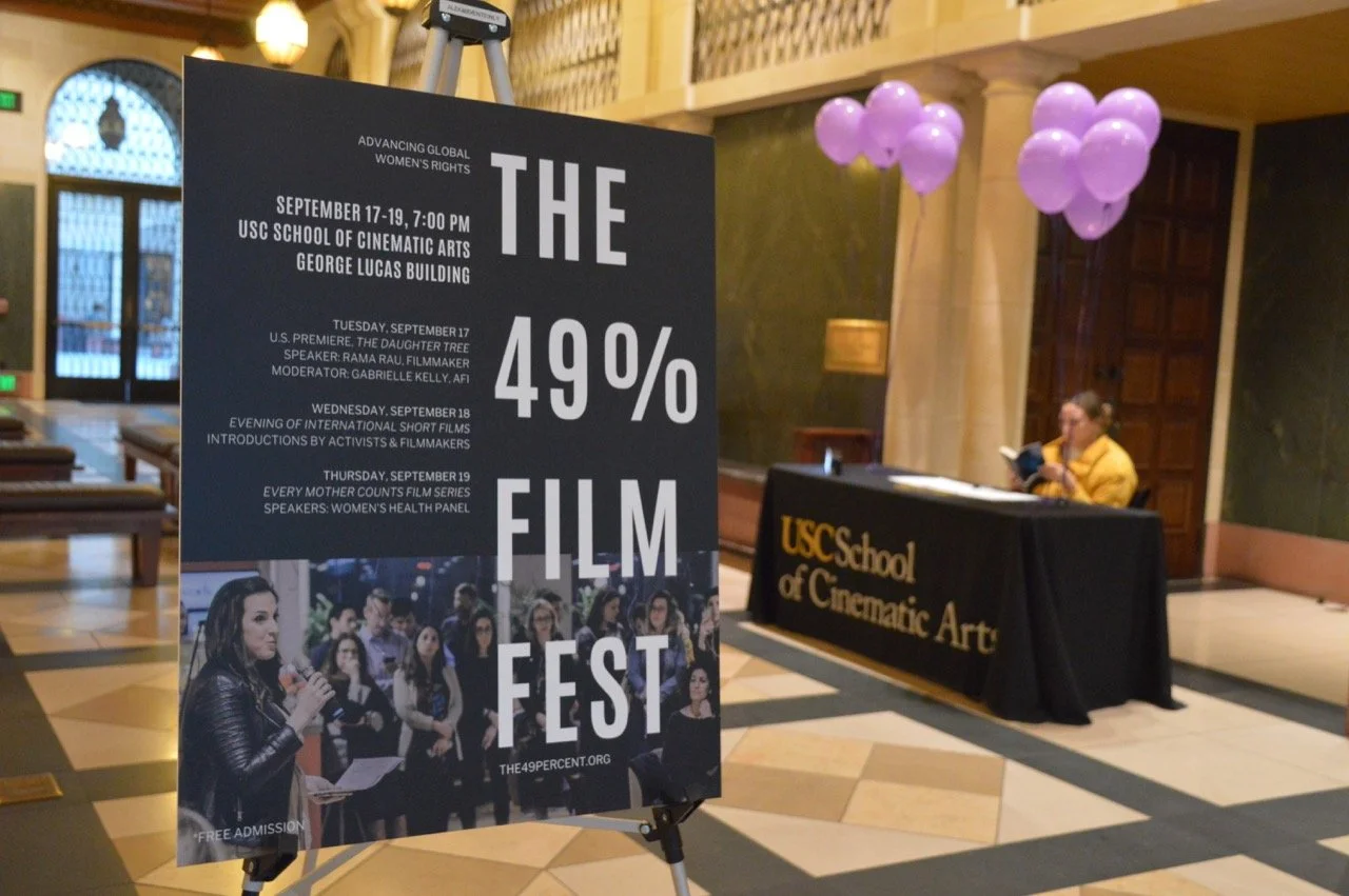 Lobby area with a poster for "The 49% Film Fest" at the USC School of Cinematic Arts, featuring event details, and a table with a banner for the school and balloons.