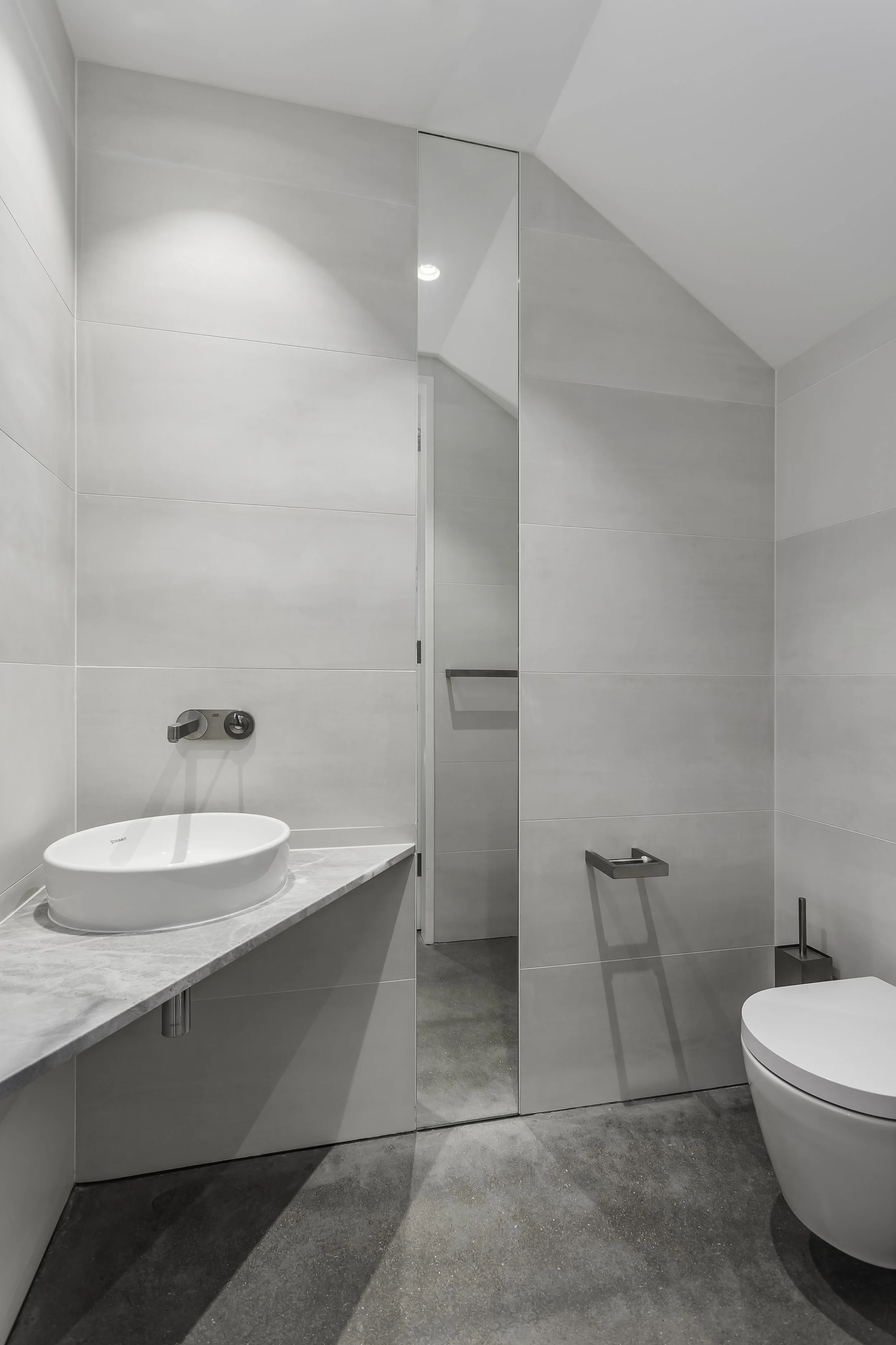 Modern minimalist bathroom with white walls, concrete floor, oval sink on marble countertop, wall-mounted faucet, and a toilet with a sleek design.