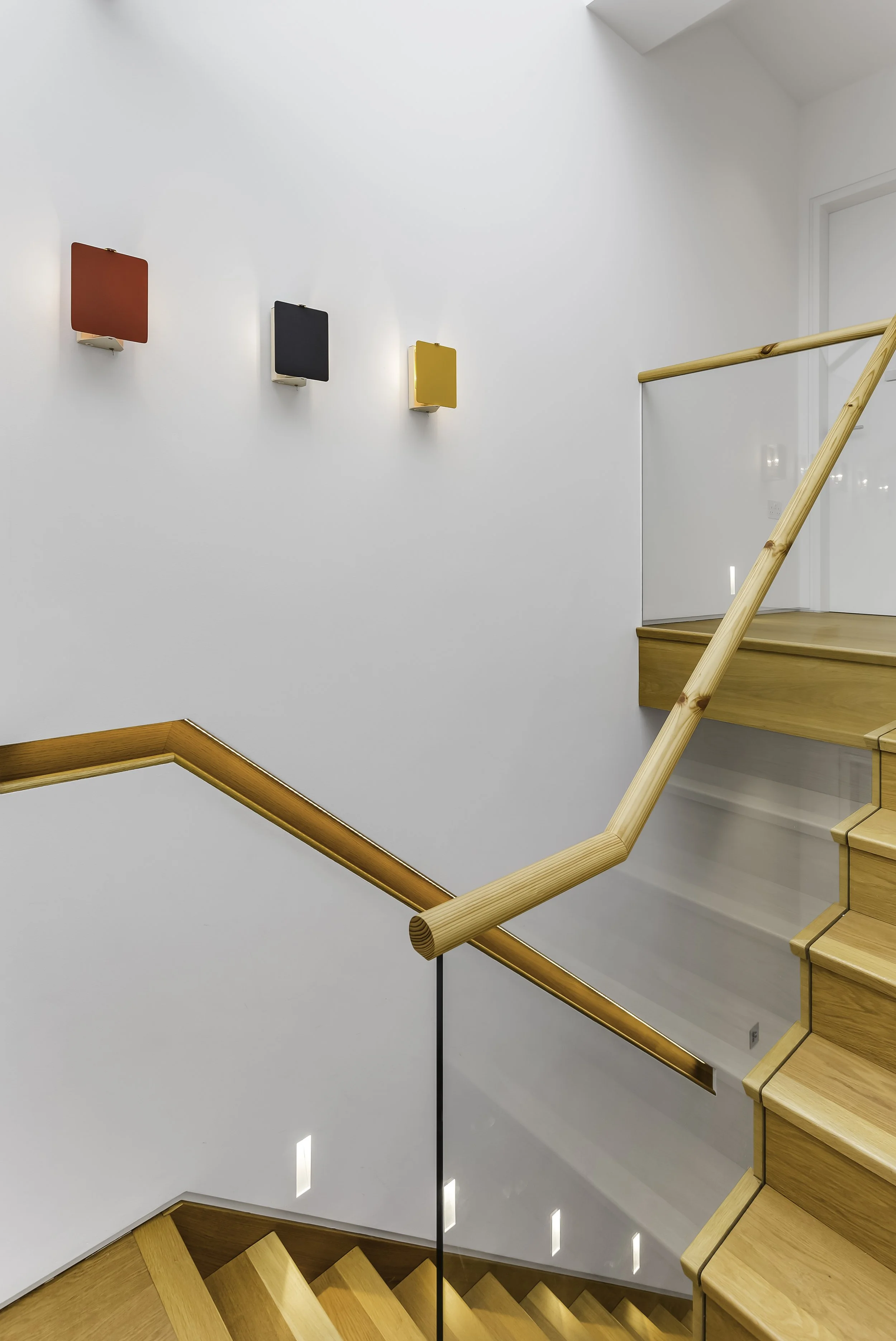 A staircase with wooden steps and a curved wooden handrail, white walls, three small colorful wall lights in red, black, and yellow, and a glass panel at the top of the stairs.