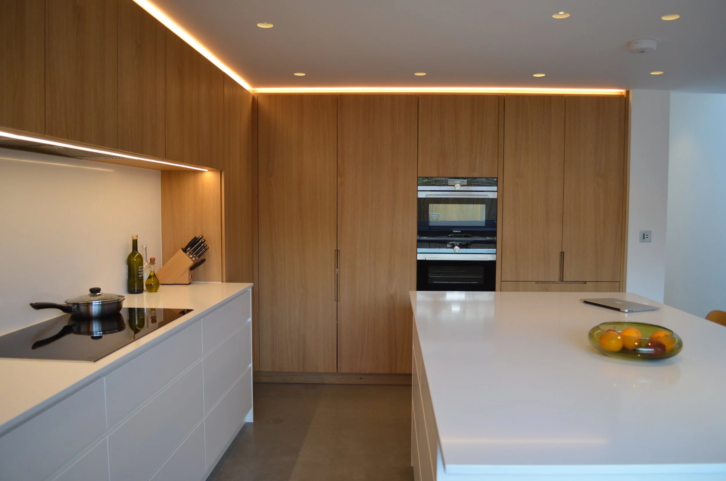 Bespoke modern kitchen with white island, wooden cabinets, and built-in appliances, illuminated by ambient lighting