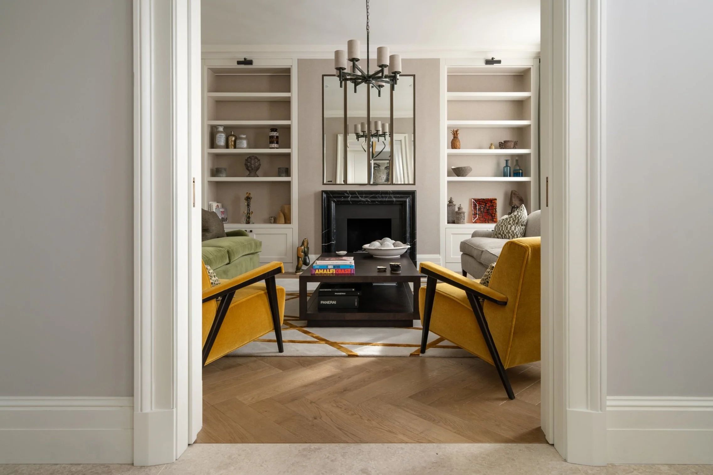 Quartet Architecture Ltd — Living room seen through doorway, with yellow armchairs, sofa, black fireplace, built-in shelves, and chandelier.