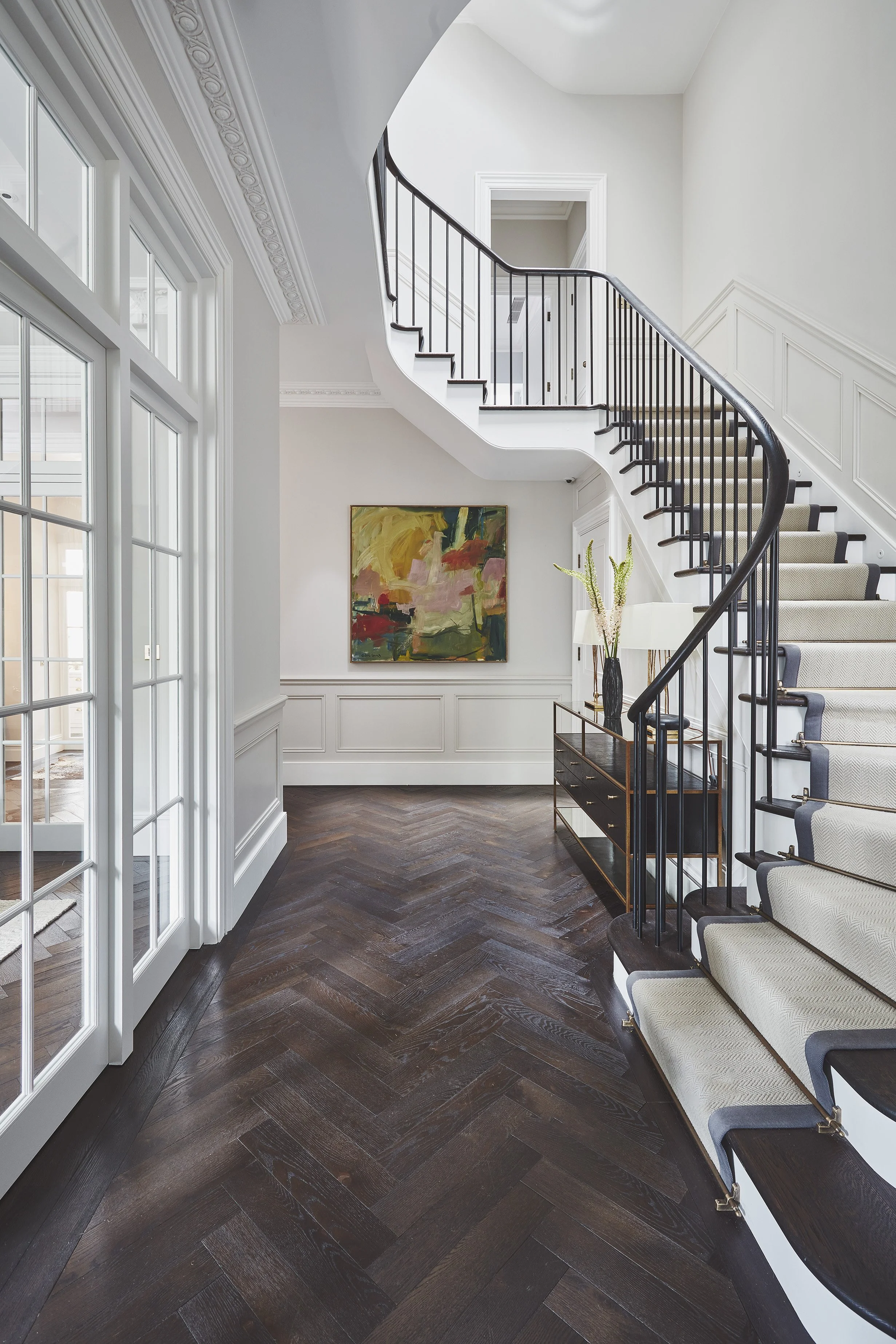 Quartet Architecture Ltd — Interior view of a modern home foyer featuring a dark wood herringbone parquet floor, a curved staircase with black metal railing and beige carpeted s