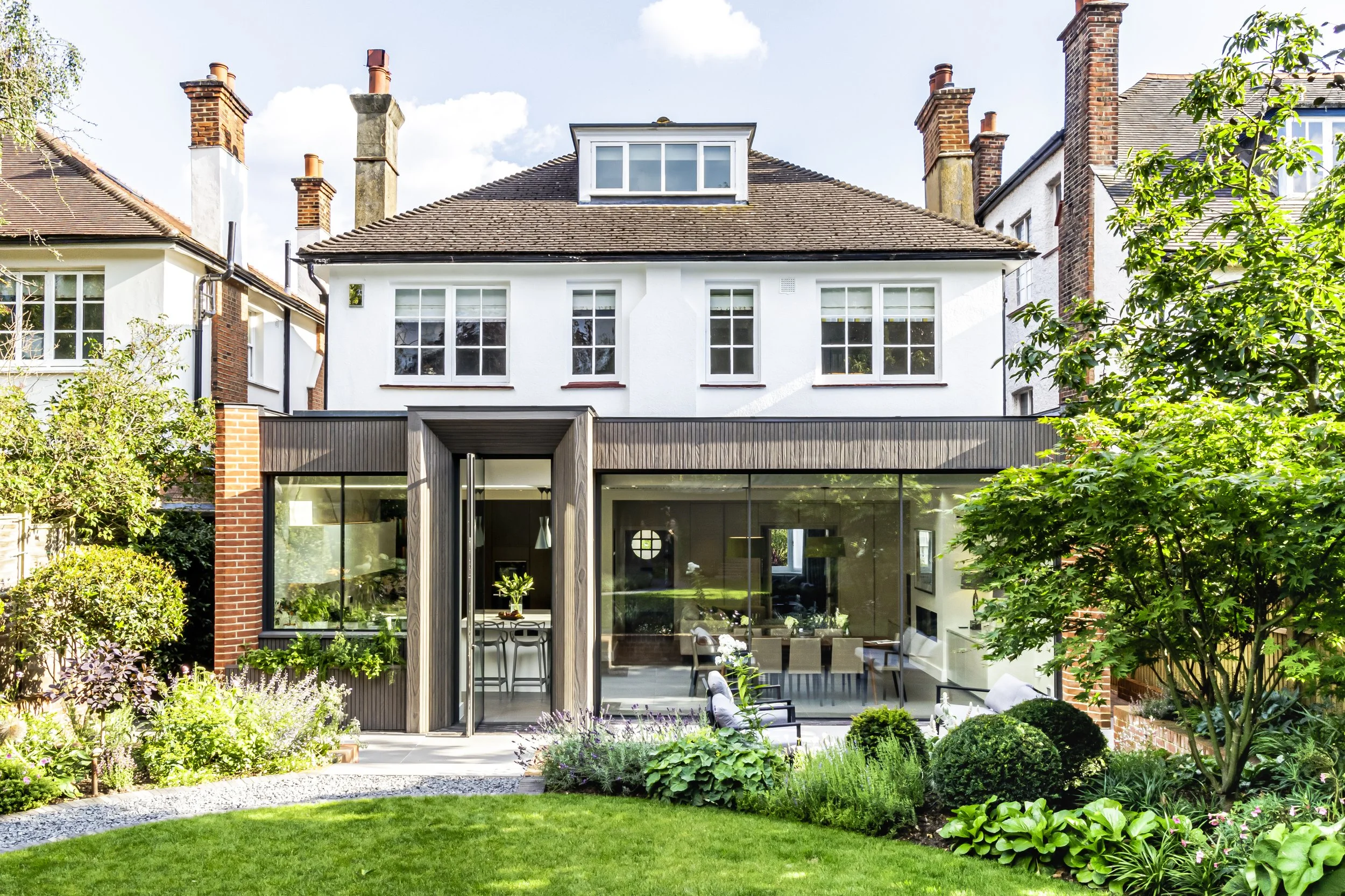 Quartet Architecture Ltd — Modern house with white upper floors and a glass rear extension surrounded by a lush green garden with trees, shrubs, and flowers.