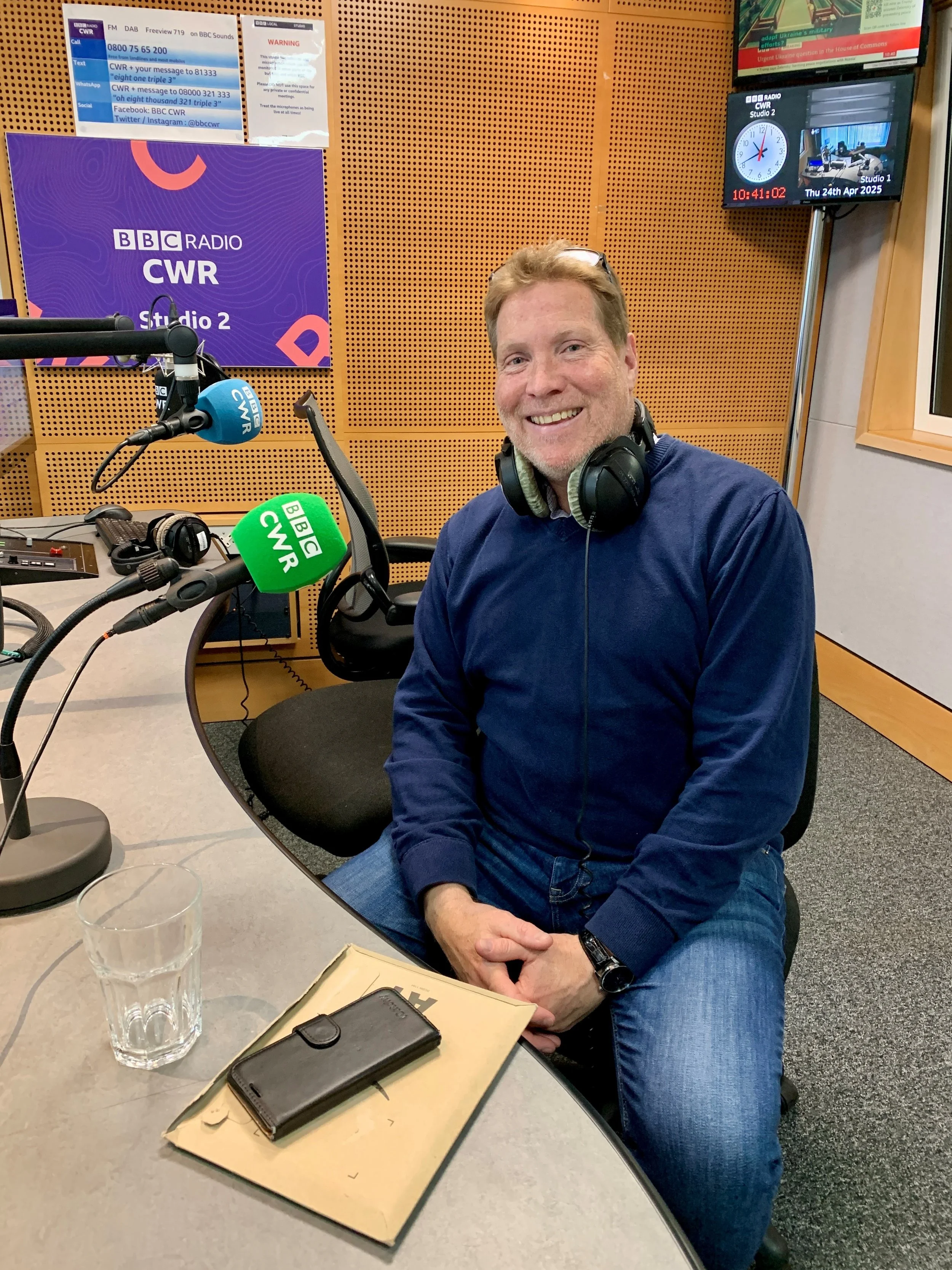 Man sitting in a radio studio with headphones around his neck, smiling at the camera. The studio has wooden panels, a sign for BBC Radio CWR Studio 2, and a clock displaying 10:41 AM on April 24, 2025. There is a microphone with a green BBC CWR foam 