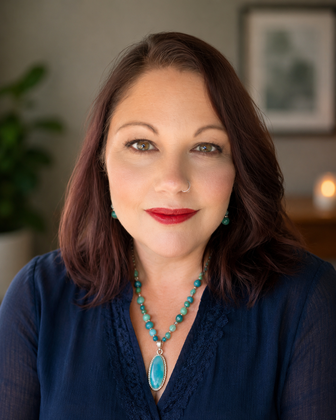 A woman with shoulder-length brown hair and light green eyes, wearing red lipstick and a nose ring, dressed in a dark blue blouse with turquoise jewelry, smiling in a warmly lit room.