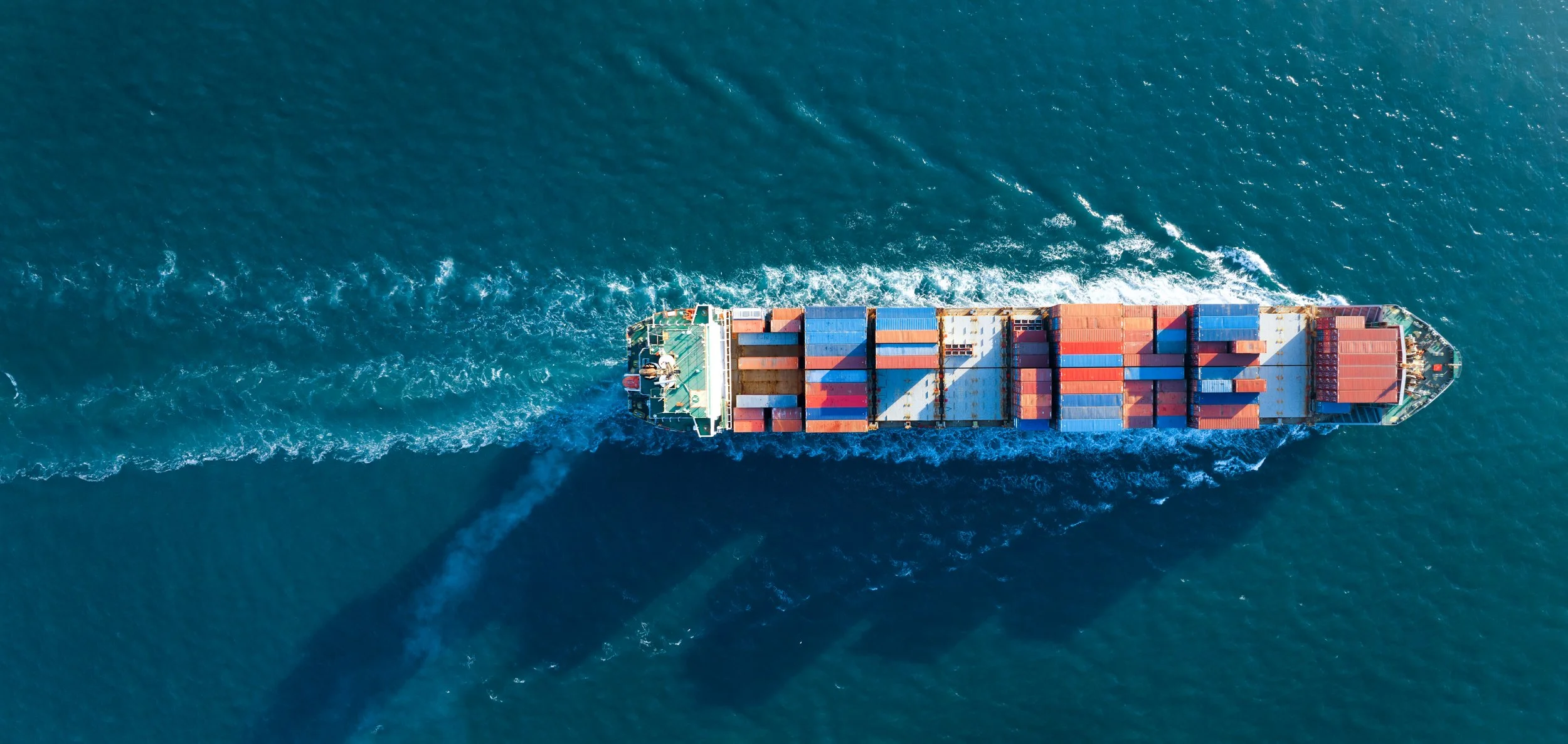 An aerial view of a cargo ship sailing through the ocean, leaving a wake behind.