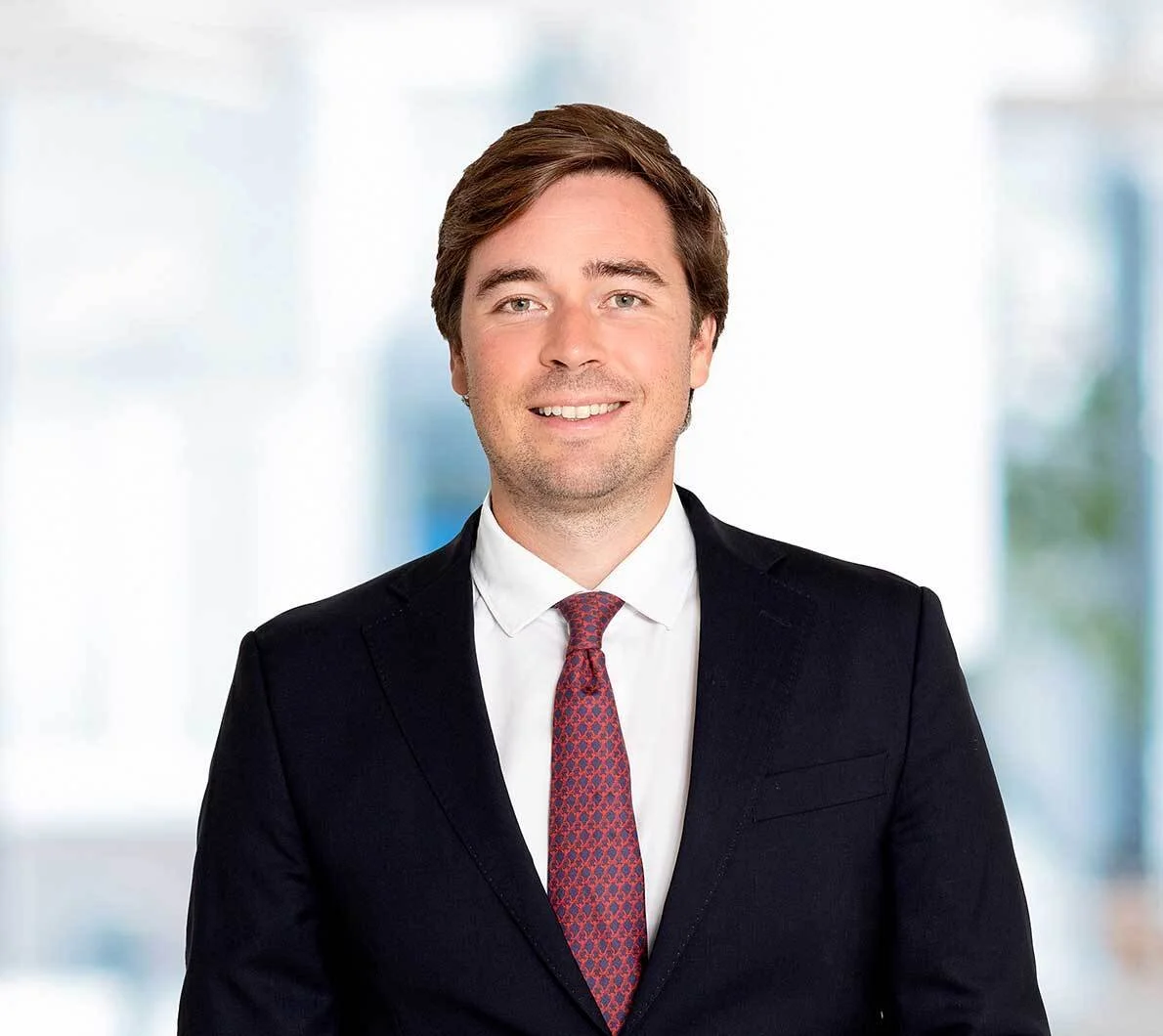 Professional headshot of a young man with brown hair, wearing a dark suit, white shirt, and patterned red tie, standing in a bright office setting.