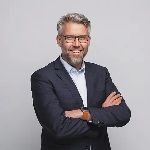 A middle-aged man with gray hair and a beard, wearing glasses, a navy blazer, and a light blue shirt, smiling and crossing his arms against a plain gray background.