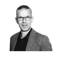 Black and white portrait of a man wearing glasses, a collared shirt, and a blazer, smiling at the camera.
