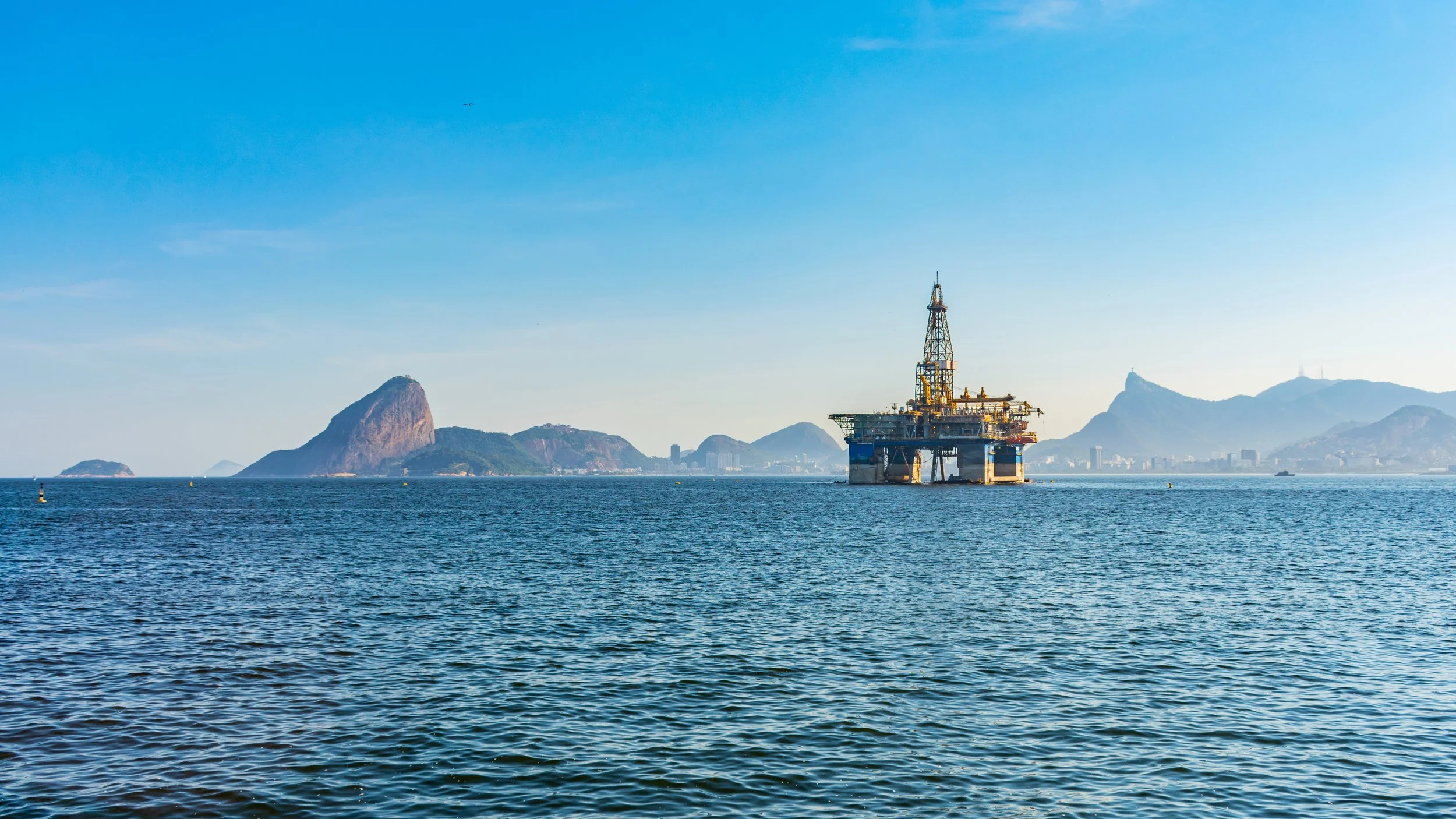 Offshore oil rig in the water with mountains and a city skyline in the background under a clear blue sky.