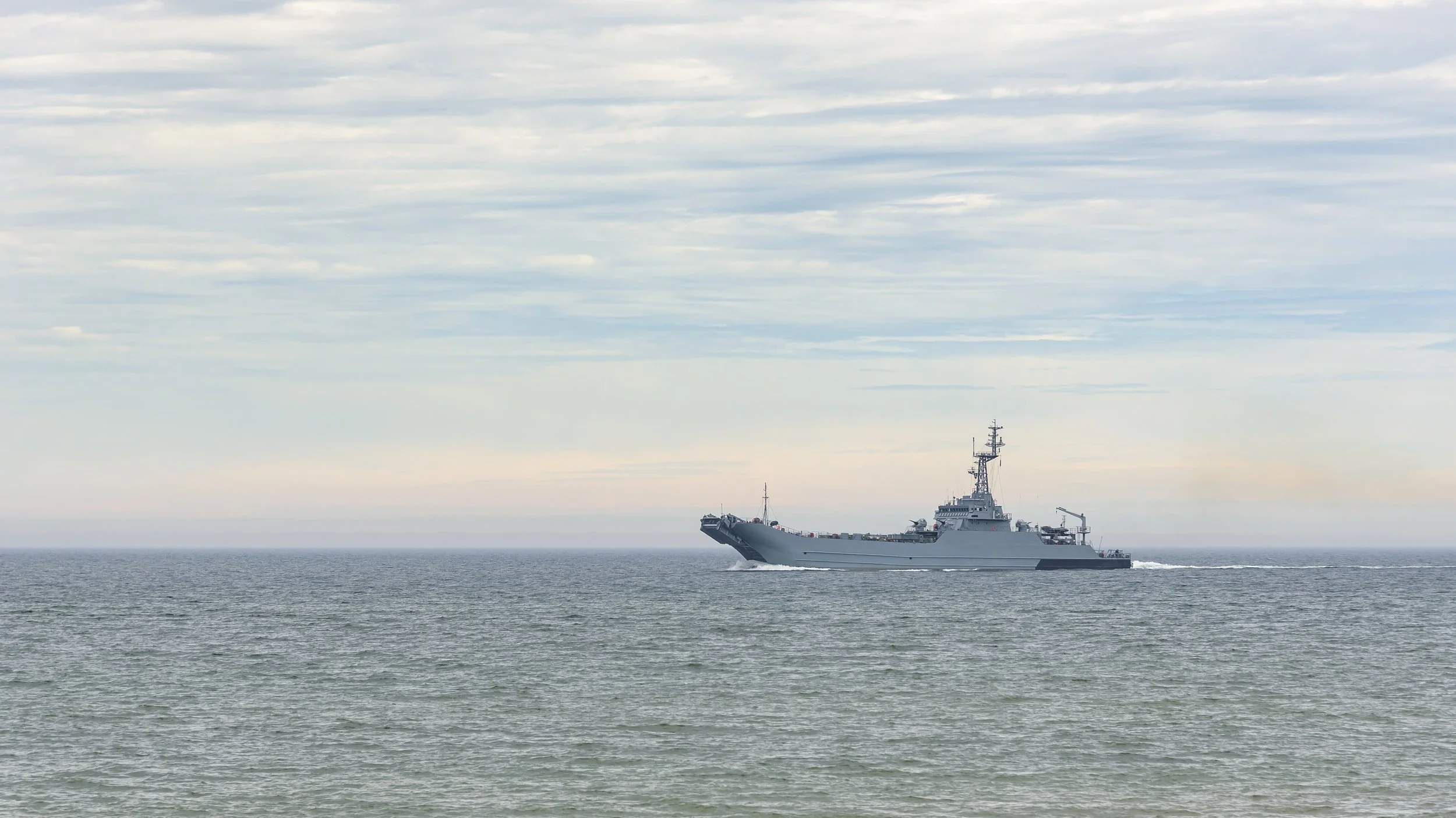 A large gray naval ship sailing on calm ocean waters under an overcast sky.